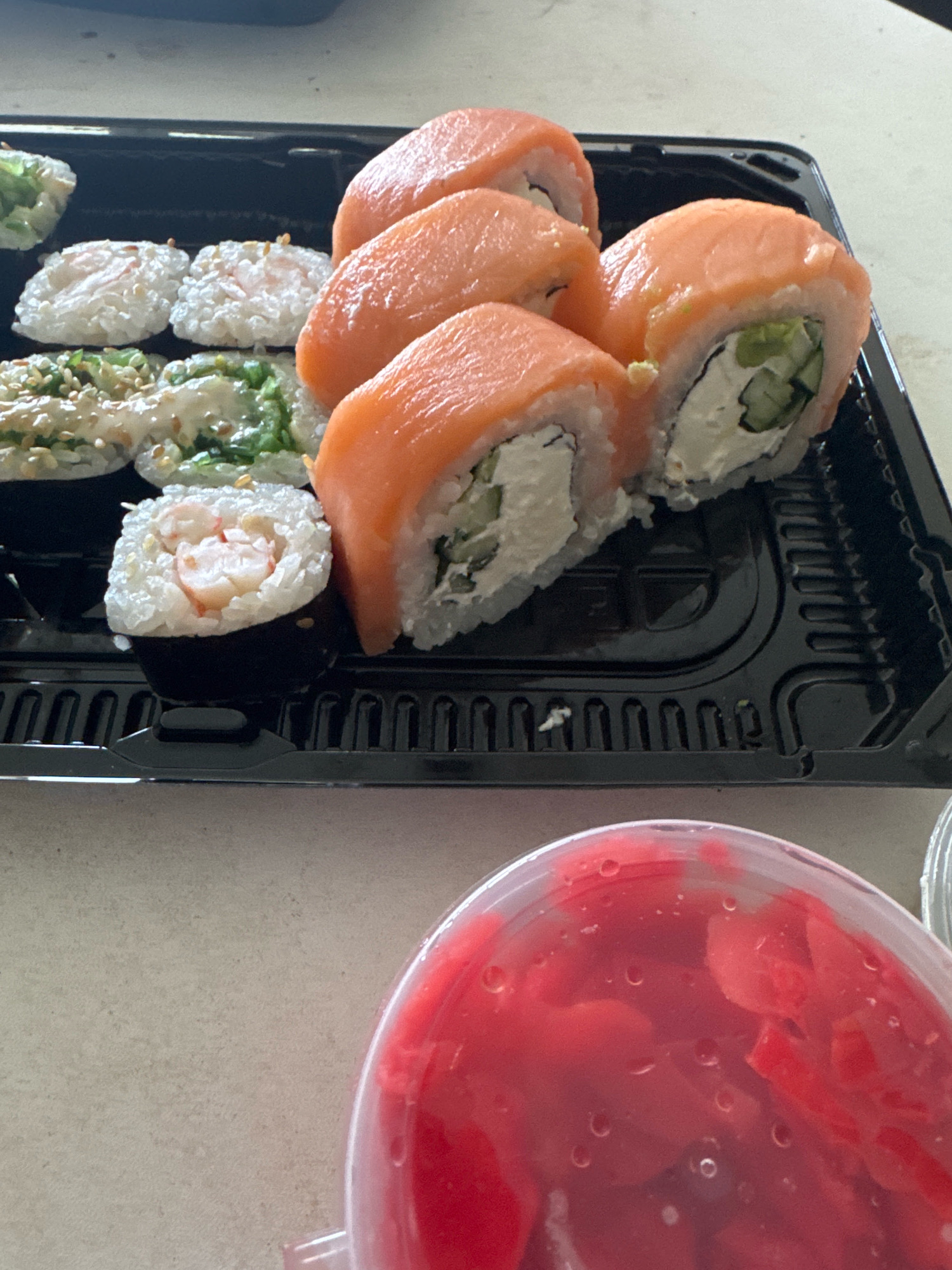 A black plastic container holds several pieces of sushi. The sushi on the right is made with salmon and cream cheese, with cucumber visible inside. To the left of the salmon rolls are smaller sushi pieces. One piece visible appears to be shrimp sushi, wrapped in nori and topped with rice and sesame seeds. Another piece has green ingredients and sesame seeds. In the foreground, a clear plastic container filled with pink pickled ginger is visible. The sushi is arranged in a black takeout container.