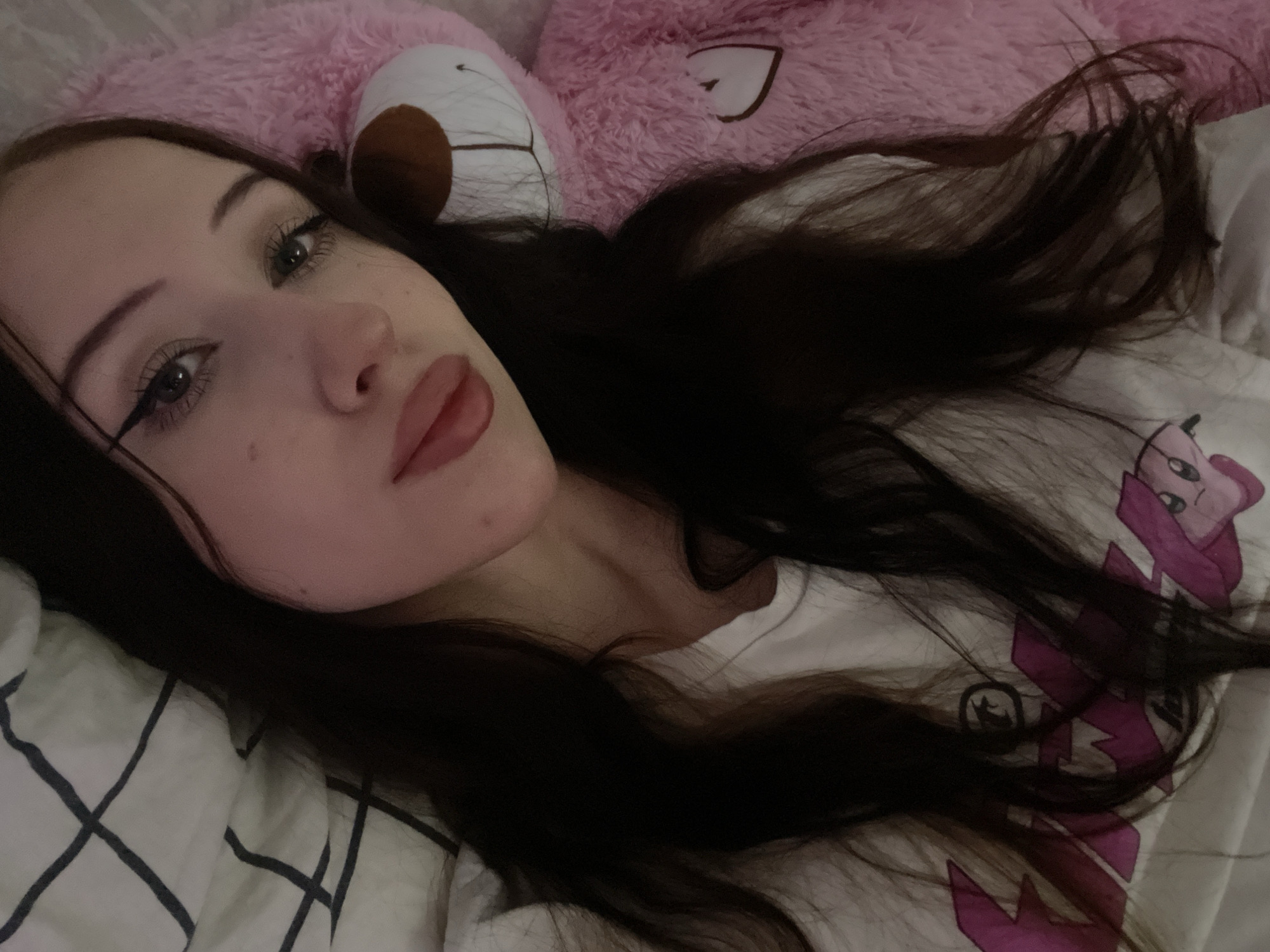The image shows a close-up, overhead shot of a young woman with long, dark hair, lying on her side. She is wearing a white t-shirt with a pink graphic design. To her right, a large pink stuffed teddy bear is visible. The background consists of a white and black checkered blanket. The scene appears to be indoors, possibly a bedroom. No specific location in Votkinsk, Russia, can be identified from the image.