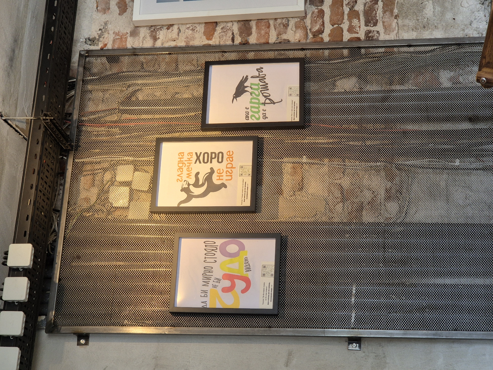 The image shows a close-up of a wall in what appears to be a cafe or restaurant, likely in Sofia, Bulgaria, given the Cyrillic text on the posters. The wall is covered by a large, industrial-style metal mesh screen. Behind the screen, cables and wires are visible, suggesting utility infrastructure. Mounted on the screen are three framed posters.
The poster on the left features colorful text in Cyrillic, with large, stylized letters spelling out "чудо" (chudo - miracle). Above it, the phrase "ДА БИ МИРНО СТОЯЛО НЕ БИ" (Da bi mirno stoyalo ne bi - If it were to stand peacefully, it wouldn't) is written. Below "чудо," it says "ВИДЯЛО" (vidyalo - seen). At the bottom of this poster, there is a QR code and some smaller text.
The central poster has a light background with a graphic of a bear silhouette with its arms raised. Cyrillic text above the bear reads "ГЛАДНА МЕЧКА" (Gladna mechka - Hungry bear). To the right of this, in a vertical orientation, is the word "ХОРО" (khoro - a type of folk dance). Below the bear, the text "НЕ ИГРАЕ" (ne igrae - doesn't play) is visible. This poster also has a QR code and some smaller text at the bottom.
The poster on the right is framed in black and features a silhouette of a crow or raven. Cyrillic text above the bird reads "ако е" (ako e - if it is). Below the bird, the words "гарва" (garva - crow) and "да е" (da e - it is) are prominent. Below this, the word "решата" (reshata - it decides/solves) is written. This poster also contains a QR code and smaller text.
The overall environment appears to be an interior space with exposed brick on the right side of the image and concrete on the left. Industrial lighting fixtures are visible at the top of the frame. The lighting is warm, casting a glow on the metal mesh. There is a hint of a wooden chair or table in the bottom right corner.