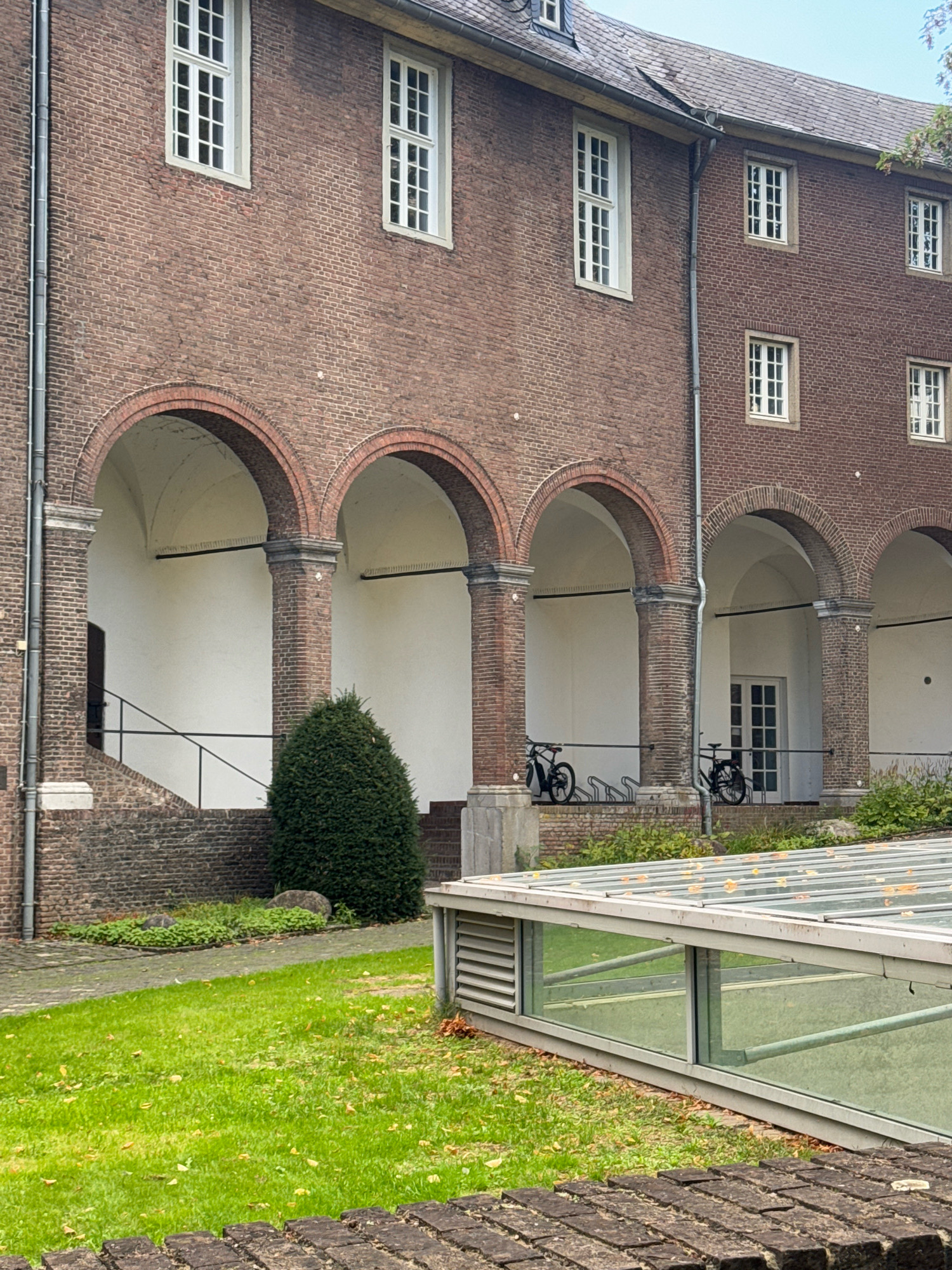 The image depicts an arched arcade along the brick facade of a building. The arcade features several brick arches supported by pillars. White walls line the interior of the arcade, and a staircase with a black railing is visible in the leftmost section. Two bicycles are parked within the arcade, one black and one dark-colored, next to a bike rack. A well-maintained green lawn with scattered yellow leaves is in the foreground, and a modern glass and metal structure, possibly a skylight or greenhouse, occupies the lower right portion of the frame. The building appears to be made of red brick and has multiple windows with white frames. The scene is set outdoors with a cloudy sky. This image is located at the Herzoghaus in Cleves, Germany, specifically the section with the arcades and the garden.