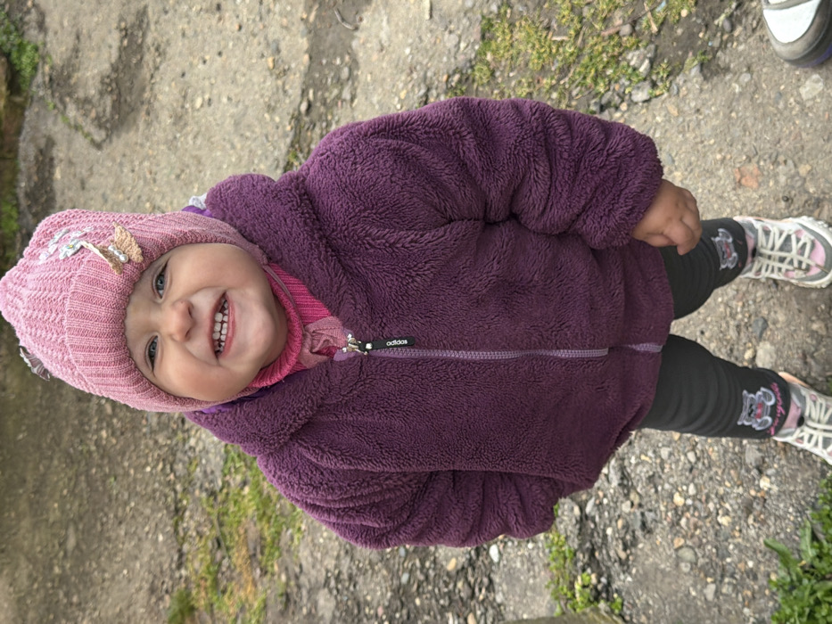 A young child stands outdoors in Komsomolskiy, Russia, captured in a full-body, high-angle view. The child, estimated to be a toddler, is smiling broadly at the camera, displaying both upper and lower teeth. They wear a pink knitted beanie adorned with light-colored decorative elements on the right side. Underneath, a pink turtleneck or high-collared shirt is visible, topped by a dark purple, long-sleesleeved, zippered fleece jacket with an "Adidas" logo on the zipper pull. Dark gray or black leggings feature cartoon animal graphics (specifically a bear on a bicycle) on each knee. The child's footwear consists of pink and white athletic shoes with gray soles. The ground beneath the child is composed of packed dirt, scattered small stones, and sparse patches of green vegetation. A portion of a gray object, possibly another shoe or a concrete slab, is visible in the lower right foreground.