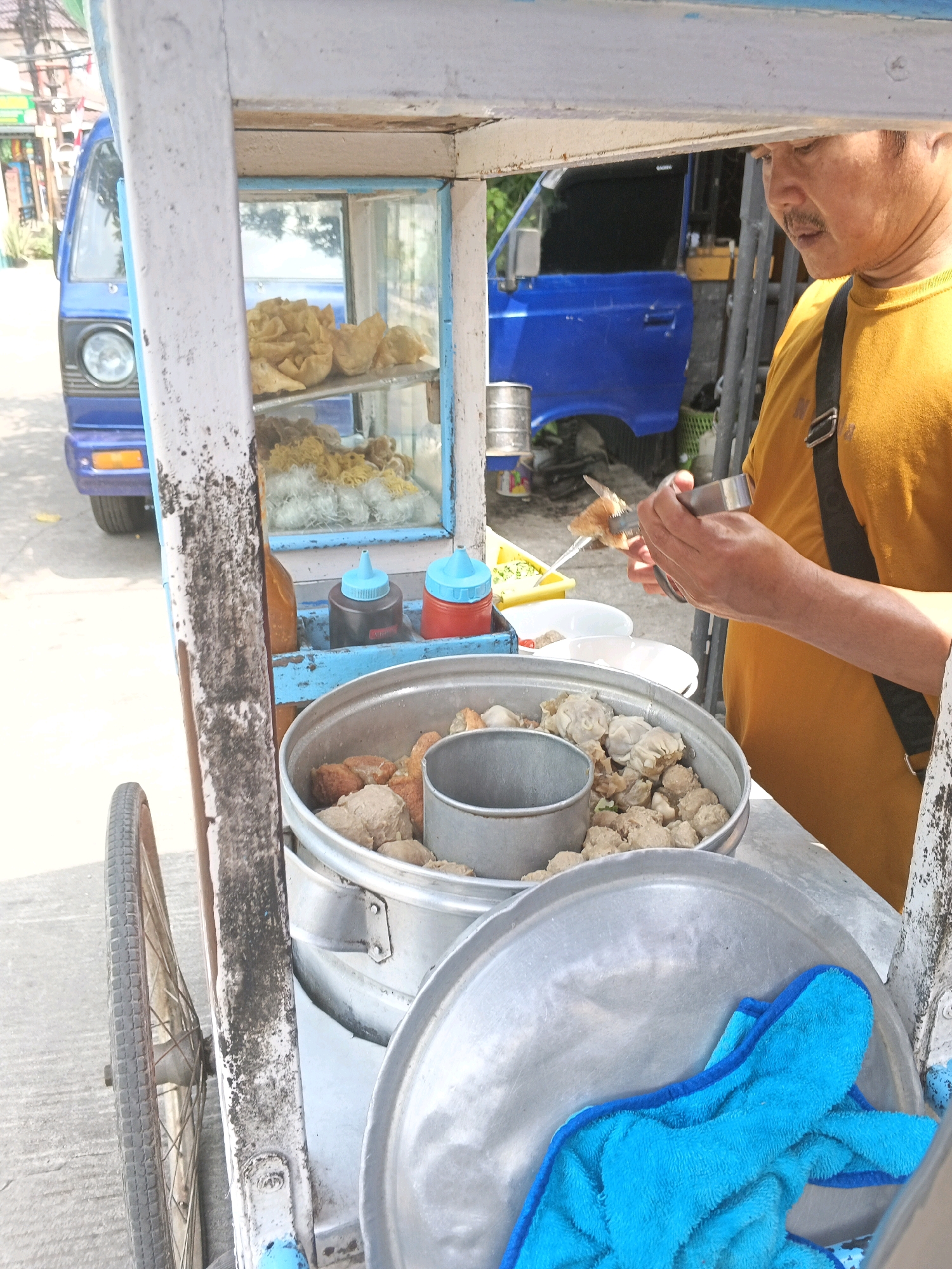The image depicts a street food vendor in Bandung City, Indonesia, preparing food from a mobile cart. The vendor, a man with a mustache wearing a yellow t-shirt, is using tongs to handle a piece of food over a metal pot filled with meatballs and other ingredients. The cart itself is made of wood and metal, painted white and blue, and has a glass display case showing fried snacks and noodles. Two condiment bottles, one red and one black with blue caps, are visible on a shelf. In the background, a blue vehicle is parked. The scene is outdoors, on a paved surface, suggesting a typical street setting in Bandung.