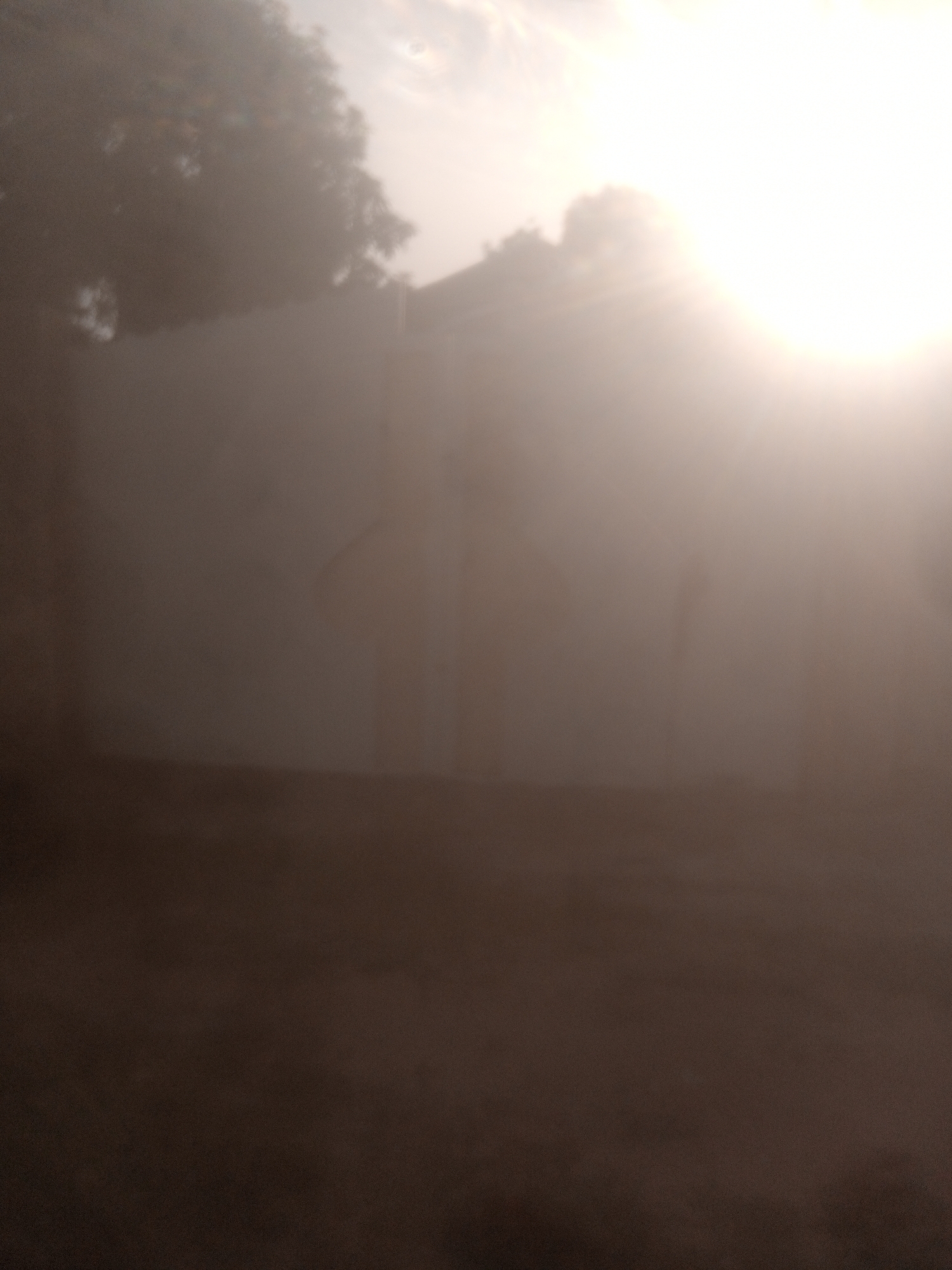 The image depicts an outdoor scene in Jalingo, Nigeria, characterized by strong backlighting from the sun, creating a hazy and diffused atmosphere. A light-colored wall, possibly a boundary or building facade, dominates the mid-ground. The upper portion of the image is obscured by the bright sun, with faint silhouettes of trees visible on the left side. The foreground is dark and appears to be unpaved ground. Due to the low light and haze, details of specific objects, people, or actions are indiscernible. The overall impression is one of a bright, possibly dusty or misty, day.