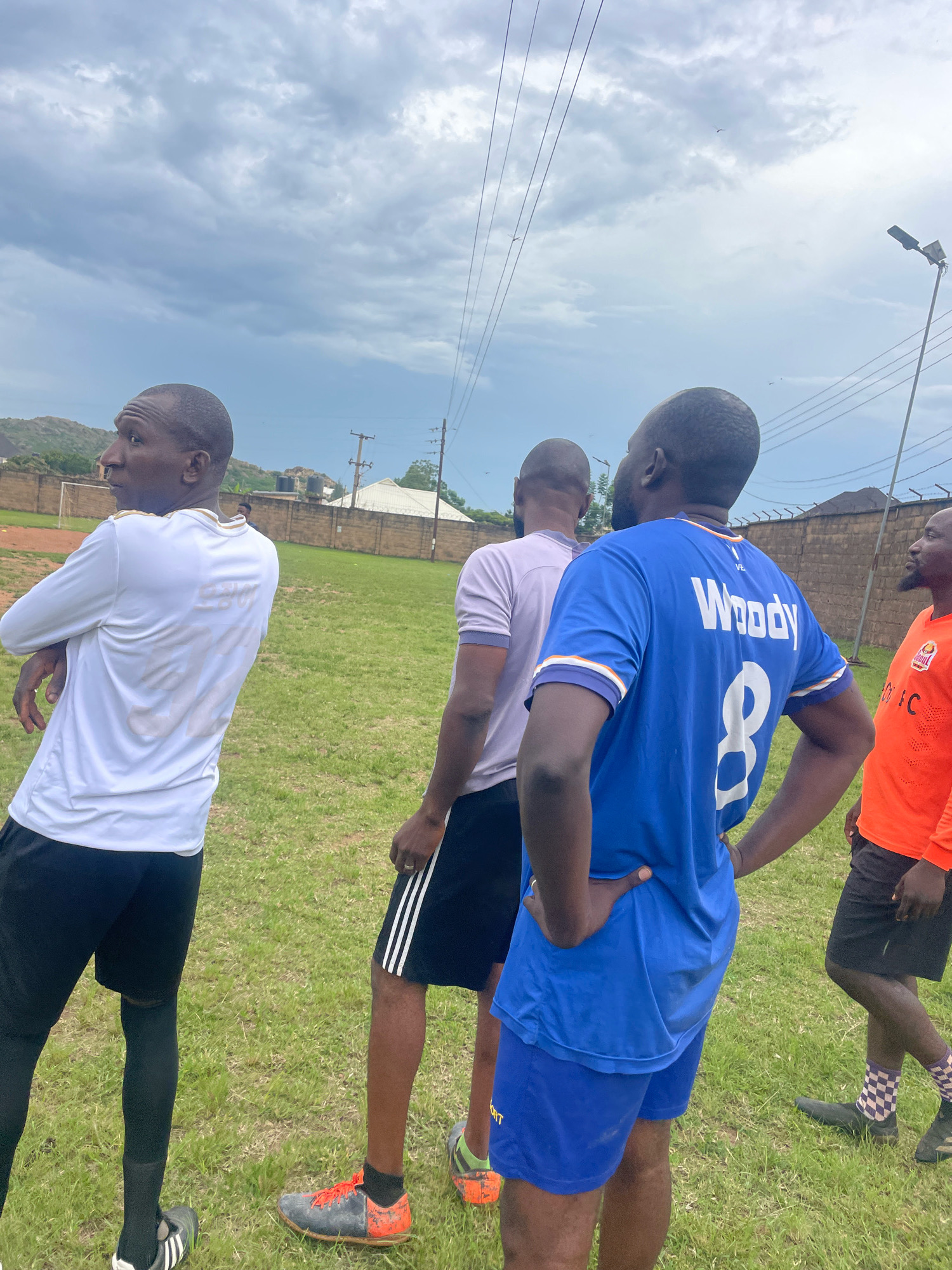 The image depicts four men on a grassy field, possibly a sports ground, under a cloudy sky. One man in the foreground, facing away from the camera, wears a bright blue soccer jersey with "Woody" and the number "8" on the back. He has his hands on his hips. To his left, another man, also facing away, wears a light purple t-shirt and black shorts with white stripes. To the left of him, a man in a white long-sleeved shirt and black shorts faces left, looking towards the side. His back is to the camera. To the right of the man in the blue jersey, a fourth man in an orange long-sleeved shirt faces right, looking away from the camera. The environment appears to be an outdoor sports facility, with a fence and some buildings visible in the background, suggesting an urban or semi-urban setting. The terrain is predominantly green grass.