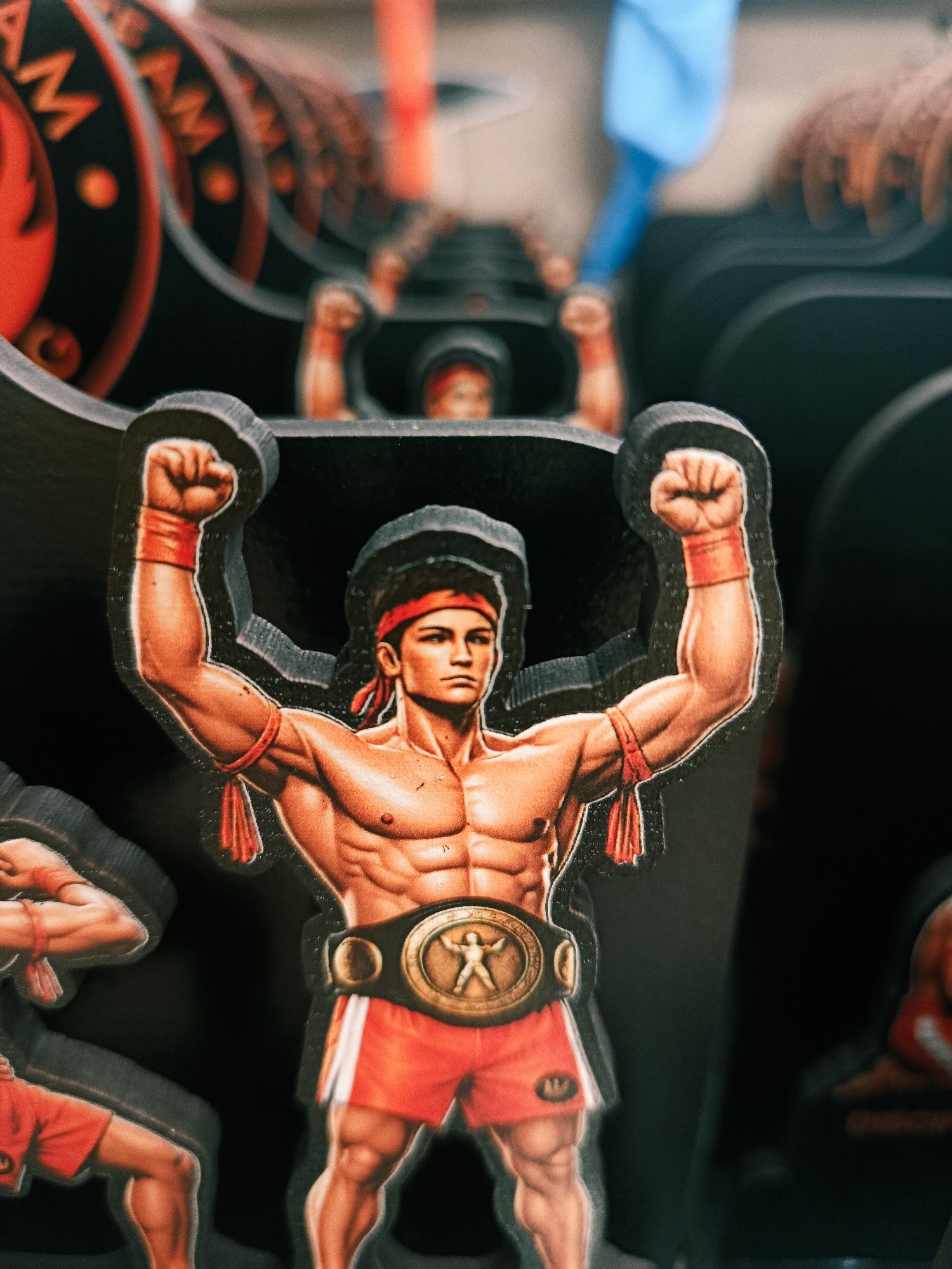 The image is a close-up, vertical, eye-level shot of a cutout of a muscular, shirtless man depicted in a fighting stance with his arms raised and fists clenched. He is wearing a red headband, red wristbands, red shorts with white trim, and a gold championship belt. The cutout has a black border and appears to be made of thick cardstock or similar material. Behind the main subject, there are several other cutouts of similar figures, creating a layered or tiered effect, and some of these are blurred due to a shallow depth of field. The background is dark, with some out-of-focus elements that suggest a display or exhibit. The lighting is artificial and highlights the textured surface of the cutouts. The location is unknown, and the image does not depict any discernible city or environmental features.