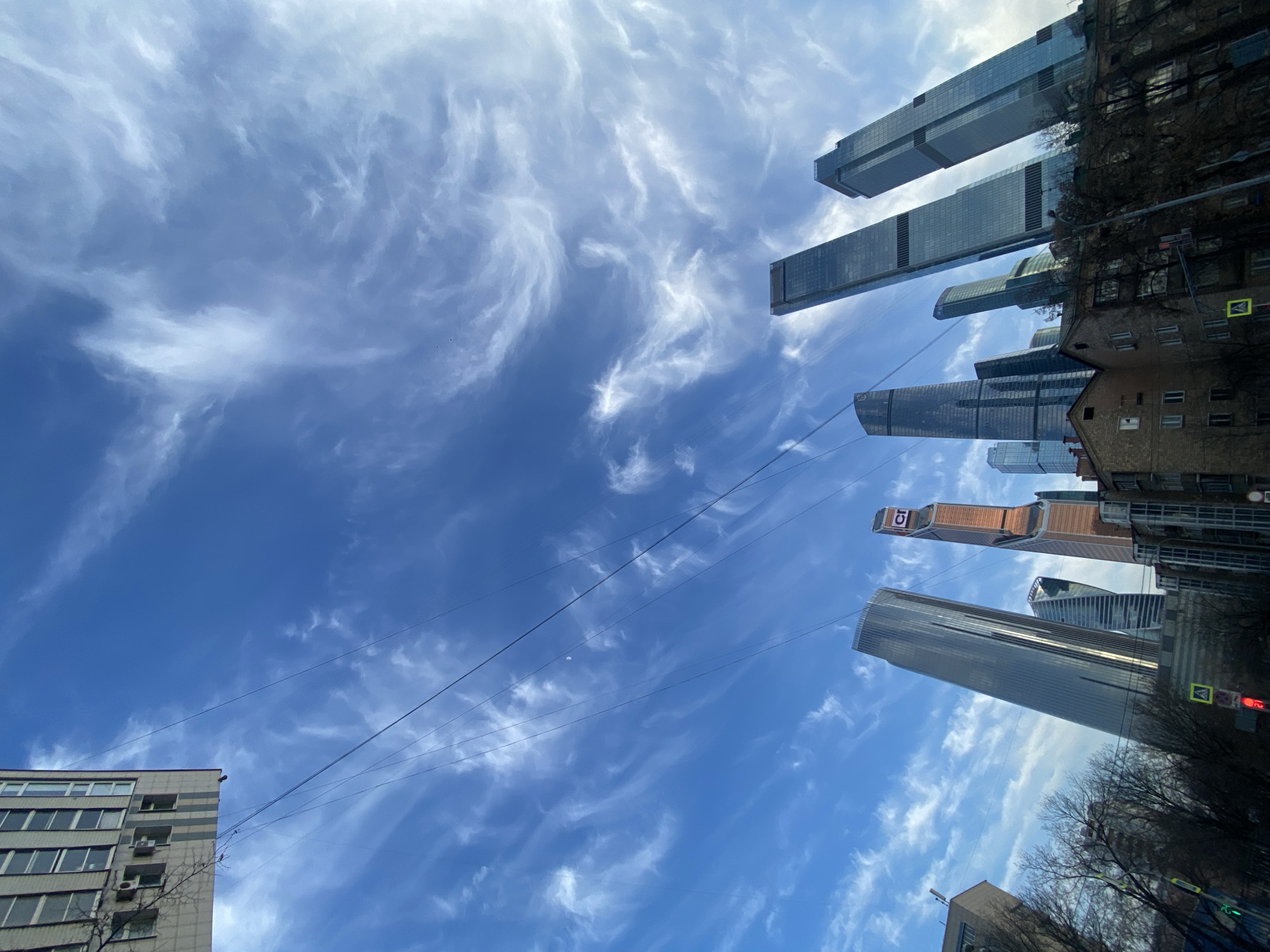 The image depicts an urban environment in Khimki, Russia, captured with a wide-angle, low-angle perspective. The scene is dominated by a bright blue sky streaked with extensive, wispy white cirrus clouds. Multiple modern skyscrapers, constructed primarily of glass and steel, occupy the central and right portions of the mid-ground, appearing to lean inwards due to lens distortion. One prominent tower features a distinct copper-colored upper section and a visible "CT" logo; another is a tall, slender glass structure. These skyscrapers are visually consistent with those found in the Moscow International Business Center. In stark contrast, older, multi-story brick and concrete buildings are visible in the foreground and lower left, their facades showing numerous windows. Bare tree branches are interspersed throughout the lower frame. Overhead power lines crisscross the sky. A faint crescent moon is visible in the upper-left quadrant. Ground-level details include a yellow pedestrian crossing sign and a red traffic light. No individuals are present. The scene is brightly illuminated by daylight, suggesting a clear day in a colder season due to the deciduous trees lacking foliage.