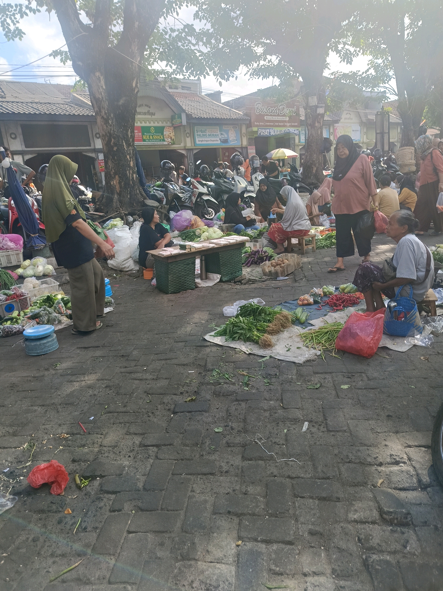 The image depicts a bustling street market scene in Yogyakarta, Indonesia, likely a traditional produce market based on the abundance of vegetables and the presence of vendors. Several individuals, predominantly women wearing headscarves and traditional Indonesian attire, are engaged in selling and purchasing goods. The market is set up along a paved street, with various stalls and temporary displays of produce on the ground, covered by tarps or mats.
In the foreground, a woman in a green hijab stands near a display of cabbages and other vegetables. To her right, a woman in a black outfit sits behind a small table laden with produce. Further to the right, a woman in a pinkish-orange top is walking, carrying a plastic bag. Another woman with grey hair, wearing a light blue shirt and dark patterned skirt, sits on a small stool, a blue woven bag beside her. She appears to be selling red chilies, long beans, and other vegetables laid out on a blue tarp.
The background reveals more market activity, with parked motorcycles indicating transportation for shoppers and vendors. Buildings with tiled roofs line the street, some displaying signs in Indonesian, including "Pusat Seragam" (Uniform Center) and "SABAT HELMET PALING MURAH" (Cheapest Helmet). The scene is illuminated by natural daylight, with sunlight filtering through the leaves of large trees that cast shadows on the ground, creating a dappled effect. The overall atmosphere suggests a typical day at a lively Indonesian market, filled with commerce and community interaction.