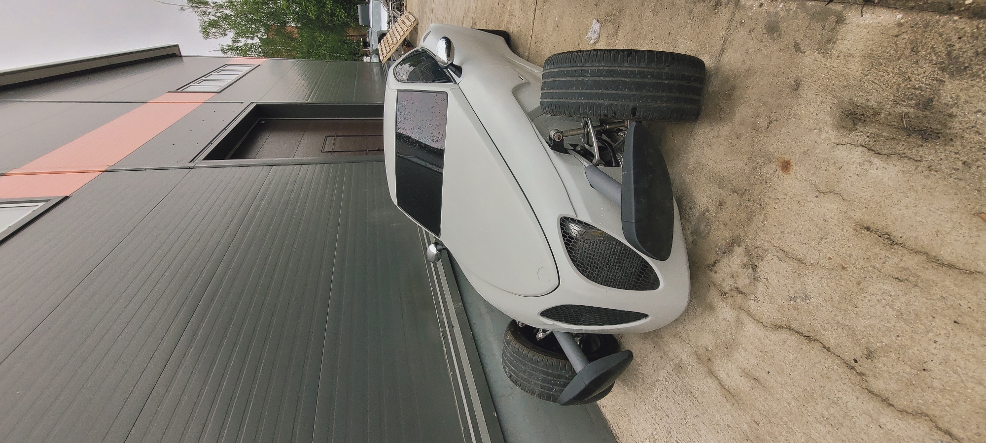 The image depicts a three-wheeled vehicle parked alongside a building. The vehicle has a white body, with black mesh grilles at the front, and its tires are black. It appears to be positioned on a concrete surface with some cracks and imperfections. The building is constructed with gray ribbed panels. It features a door, some windows, and a horizontal stripe of orange color above the door. In the background, there are some trees and possibly other vehicles. The location is in the city of Unknown, Unknown.