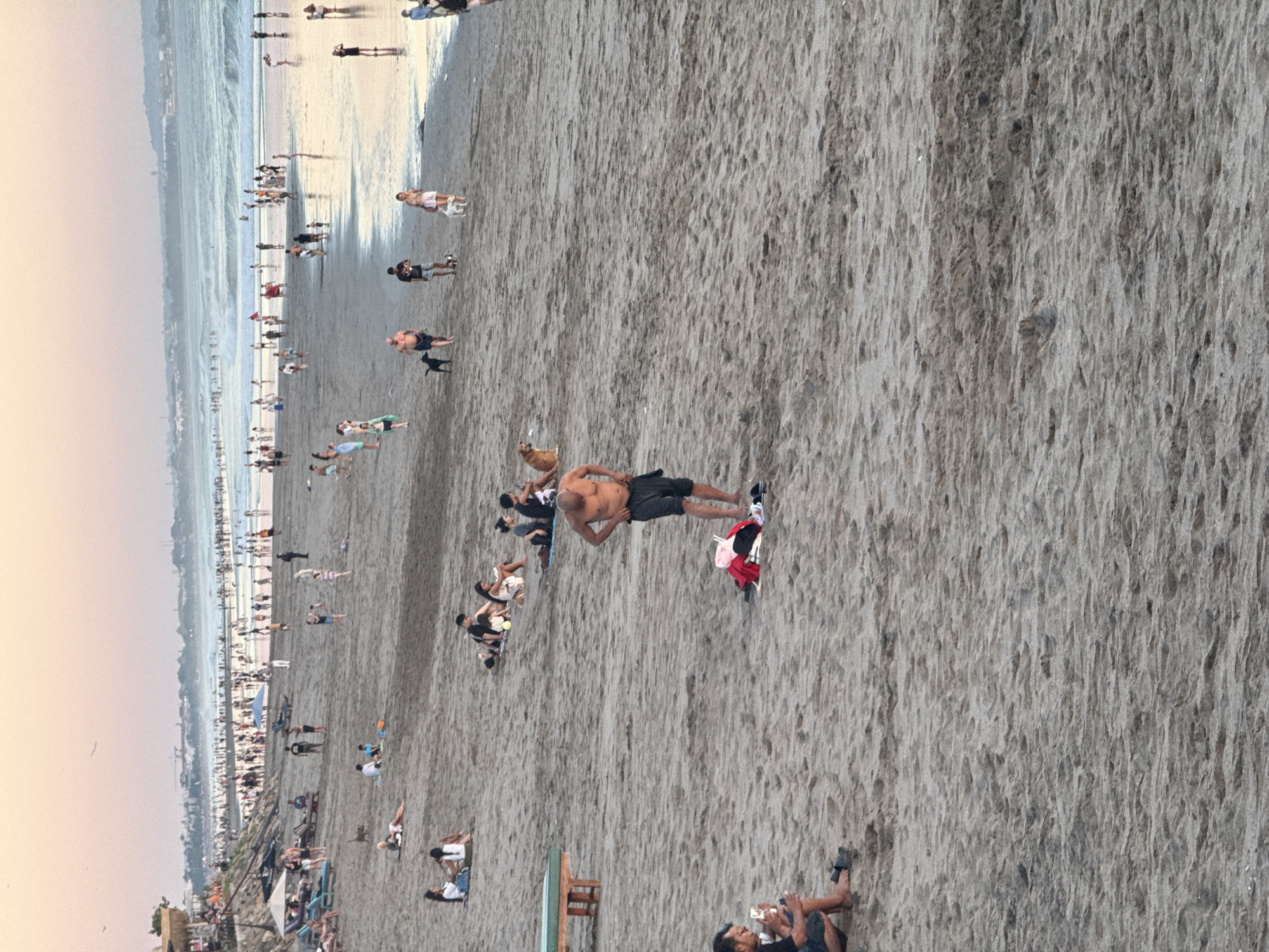 This is a full-shot, eye-level photograph of a crowded beach in North Kuta, Indonesia, during what appears to be sunset. The sky is a gradient of pale pink and orange, with the sun low on the horizon. The ocean is visible in the background, with gentle waves and numerous people wading and swimming. The beach itself is wide and sandy, with many people relaxing, walking, and playing.
In the foreground, a man stands on the sand, partially undressing and holding his shorts. Nearby, a pile of clothes, including a red shirt and a white shirt, lies on the sand, along with a pair of flip-flops. To the left, another man sits on the sand, looking at his phone.
Further up the beach, several groups of people are scattered. One man is walking a black dog. Another man is walking a small white dog. There are also people sitting on lounge chairs under umbrellas, and children playing in the sand. In the distance, the coastline is lined with buildings and palm trees. The overall atmosphere is one of a lively beach scene with many people enjoying the end of the day.