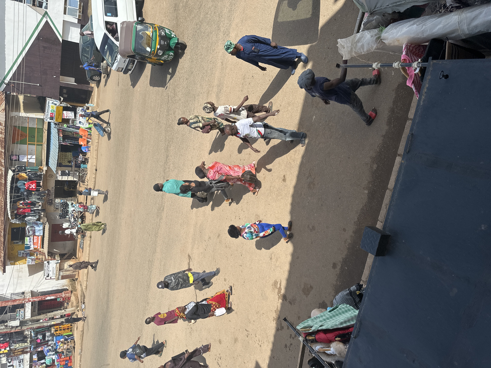 The image depicts a street scene in Bukuru, Nigeria. A diverse group of individuals are visible on a dusty road, with buildings lining one side. In the foreground, a dark, presumably metal, structure dominates the bottom portion of the frame, with various items of clothing hanging from a rack above it.
The street is populated by a number of people, some walking, some standing. A woman in a bright, multi-colored top and blue pants is walking towards the right. Further back, a man in a dark blue robe and headscarf stands looking to his left, while another man in a white polo shirt and jeans stands facing forward. To the left, a woman in a pink and red patterned dress walks with her back to the camera, carrying a white bag. Nearby, a young girl in a colorful patterned dress walks.
In the middle ground, several individuals are moving. A man in a green robe walks away from the camera, and another man in a black and camouflage jacket walks towards the left. A man in a dark shirt and pants pushes a blue trolley with wheels down the street. A white van and a green and yellow auto-rickshaw are parked on the road.
The buildings on the left appear to be shops with goods displayed outside. The environment suggests a bustling marketplace or a street with shops and residences. The lighting indicates it is daytime, with clear shadows cast on the ground.