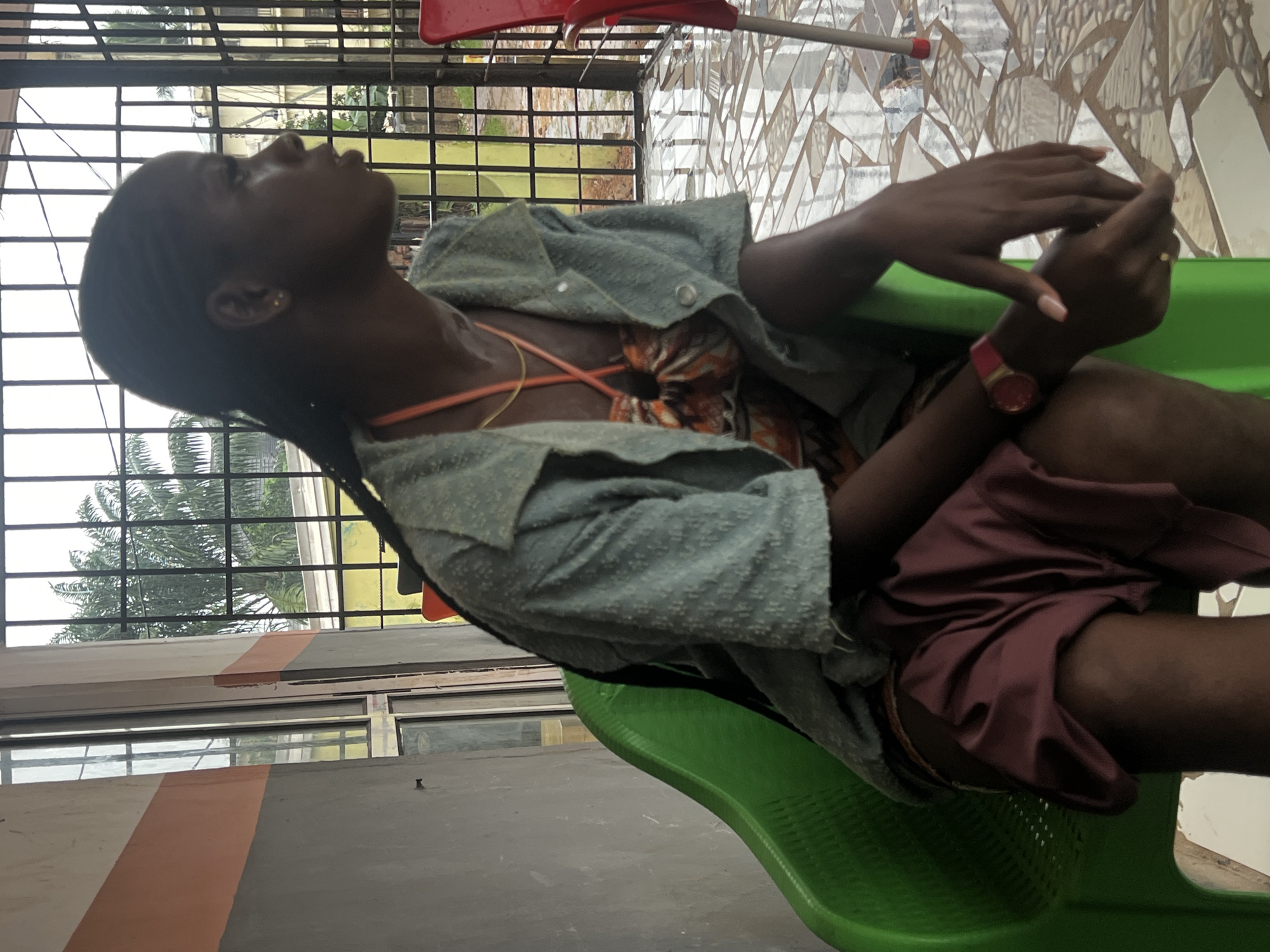 A young Black woman is seated in a green plastic chair, her body angled away from the camera, facing the right side of the frame. She has braids and is wearing a light blue, short-sleeved, button-up shirt over a patterned dress. A thin gold chain is visible around her neck, and an orange cord is also present. She wears a red watch on her left wrist. Her legs are crossed at the knee, with a maroon garment draped over her lap. Her hands are clasped in her lap. The background consists of a wall with horizontal bands of gray and orange, a large window, and a barred metal gate through which palm trees and another building are visible. The floor is tiled with a mosaic pattern. The scene appears to be an outdoor seating area, possibly at a commercial establishment or residence in Nkpor, Nigeria.
