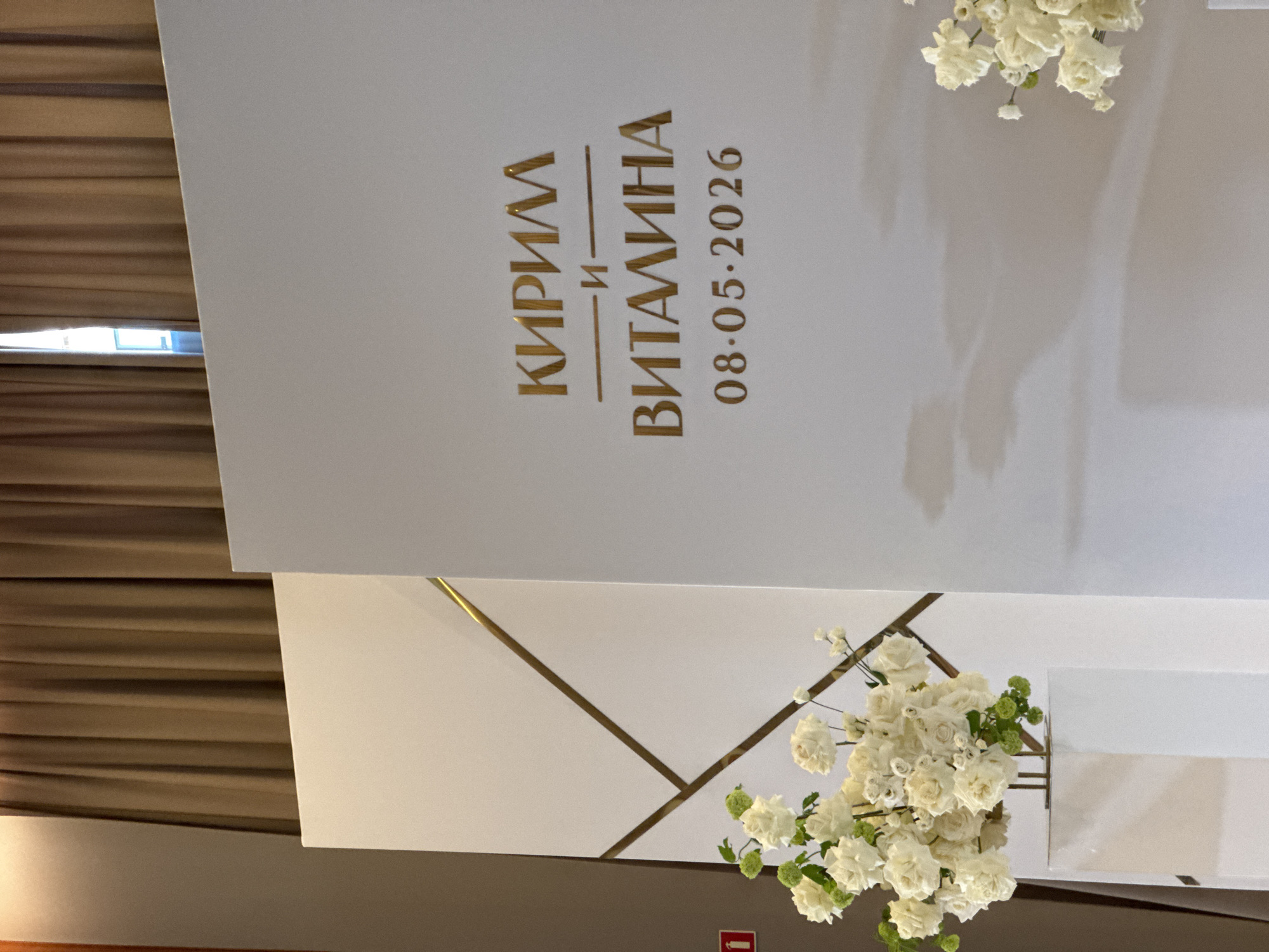 The image shows a wedding sign, likely located in Yekaterinburg, Russia, based on the Cyrillic text. The sign is white with gold lettering. The names "Кирилл" (Kirill) and "Виталина" (Vitalina) are displayed with a conjunction "и" (and) between them, indicating they are the names of the couple getting married. Below the names, the date "08.05.2026" is displayed, likely the wedding date. The background is white, with gold trim. There are white flowers in vases near the sign. A wall with brown curtains is partially visible in the background.