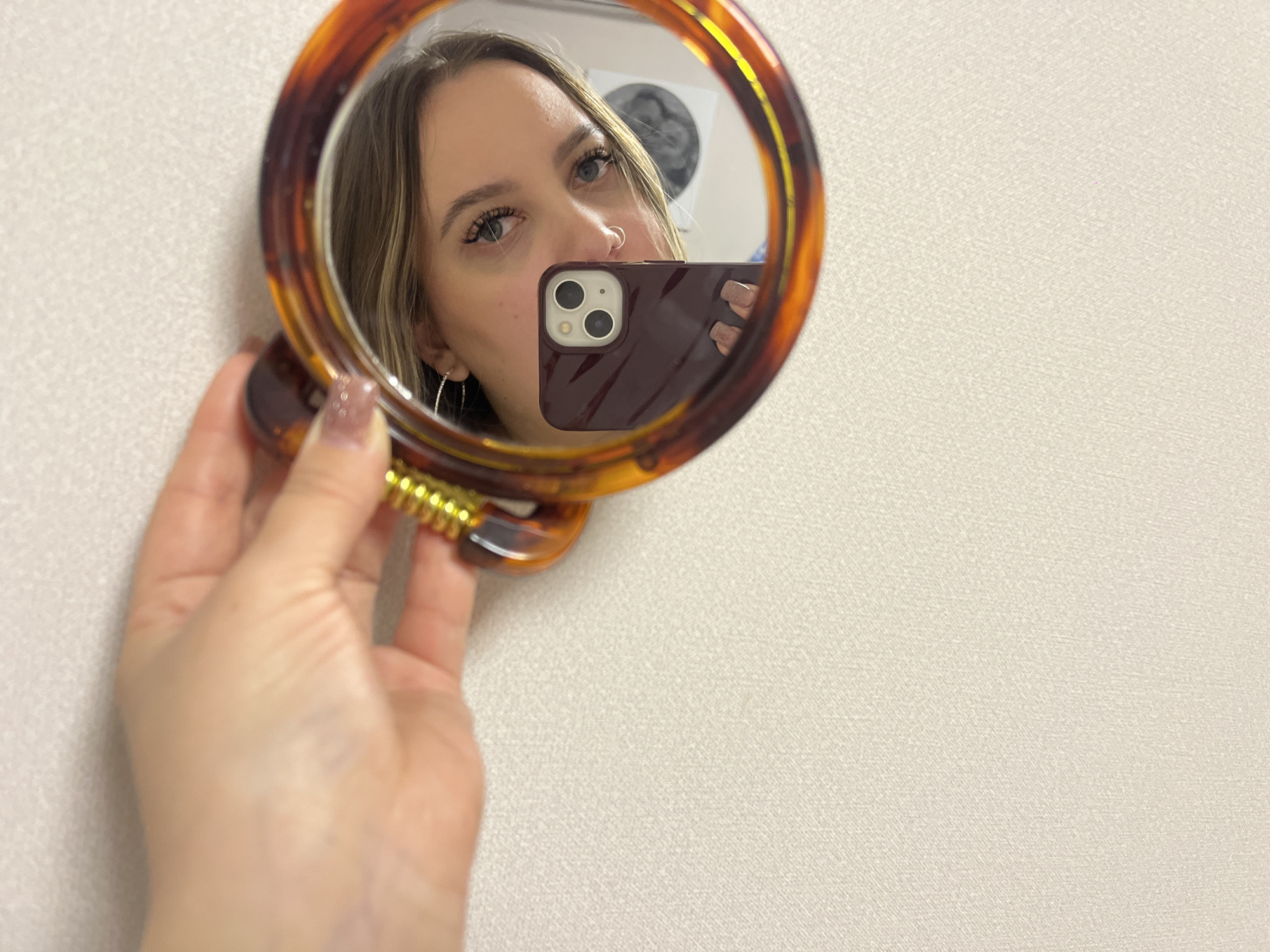 The image captures a close-up indoor shot, featuring a young woman reflected in a handheld mirror. The mirror has a distinctive tortoiseshell-patterned frame and a gold-colored, spiral-detailed handle, which she holds with her left hand, revealing nails painted with a light, shimmery polish.
Within the mirror's reflection, the woman's face is partially visible, showing her light-colored hair, a nose piercing, and hoop earrings. Her eyes are open, and she appears to be wearing mascara. She holds a dark reddish-brown smartphone, with its multiple camera lenses clearly visible, using it to photograph her reflection. The background visible in the mirror's reflection is blurry but suggests an indoor setting, possibly with a circular poster or object on a wall.
Outside the mirror, the wall serving as the immediate background is a light, neutral color with a subtle woven or textured pattern. The activity taking place is a selfie being captured through a mirror, indicating self-portraiture. The lighting appears to be ambient indoor light. No specific time of day or weather conditions are discernible. There is no visible text in the image. The photo is set in Krasnodar, Russia.