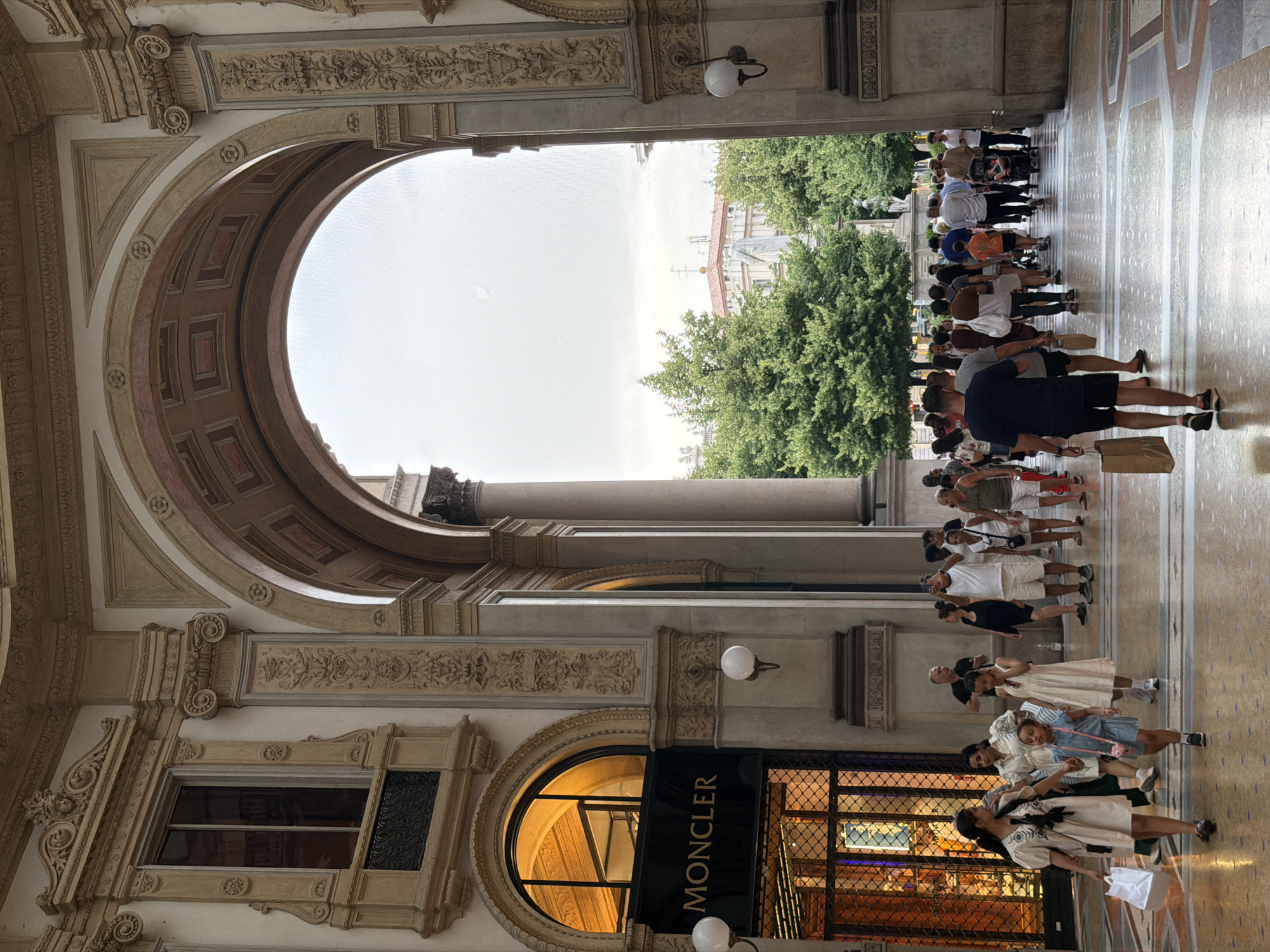 The image shows a view through a large arched entryway in Milan, Italy, likely the Galleria Vittorio Emanuele II, given its architecture and the presence of the Moncler store. The interior is characterized by ornate classical architecture with elaborate moldings and columns. Sunlight streams in from the left, illuminating a Moncler storefront with its logo prominently displayed. In the foreground and middle ground, a diverse group of people are walking, some carrying shopping bags. They are dressed in casual summer attire. Beyond the archway, lush green trees and classical buildings are visible against a bright, overcast sky. The flooring is a polished mosaic pattern.
