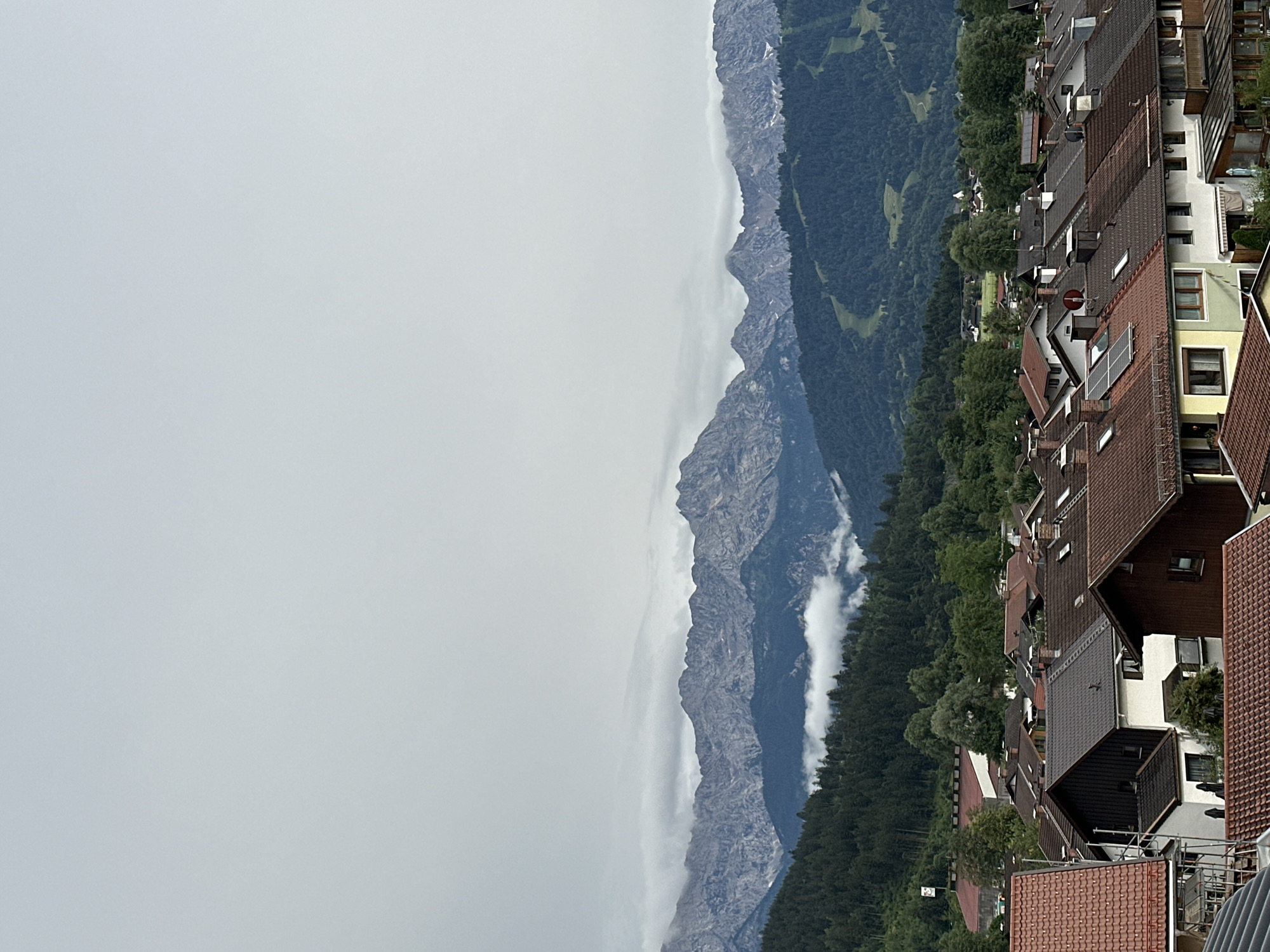 This is a vertically oriented photograph taken from a high vantage point, likely a balcony or rooftop, overlooking a town nestled against a mountain range. The scene is split between the man-made structures of the town and the natural beauty of the mountains.
In the foreground, the focus is on a dense cluster of multi-story houses with predominantly terracotta tiled roofs. Many of these houses feature dormer windows, solar panels, and satellite dishes, indicating a residential area. The architecture suggests a European setting. Behind the houses, a lush, dark green forest stretches upwards, eventually giving way to the rugged, grey peaks of the mountains. The mountains are partially obscured by a layer of white clouds that cling to their slopes, creating a sense of depth and mystery.
The weather appears overcast, with a significant portion of the sky on the left of the image being a pale, uniform grey. This suggests either early morning, late afternoon, or a generally cloudy day. The diffused light further contributes to the muted color palette of the scene. There are no people or overt activities visible, implying a quiet, perhaps residential, moment captured in time. The location is identified as Garmisch-Partenkirchen, Germany, which is known for its Alpine scenery, aligning with the mountainous backdrop and the style of the buildings. No text is visible in the image.