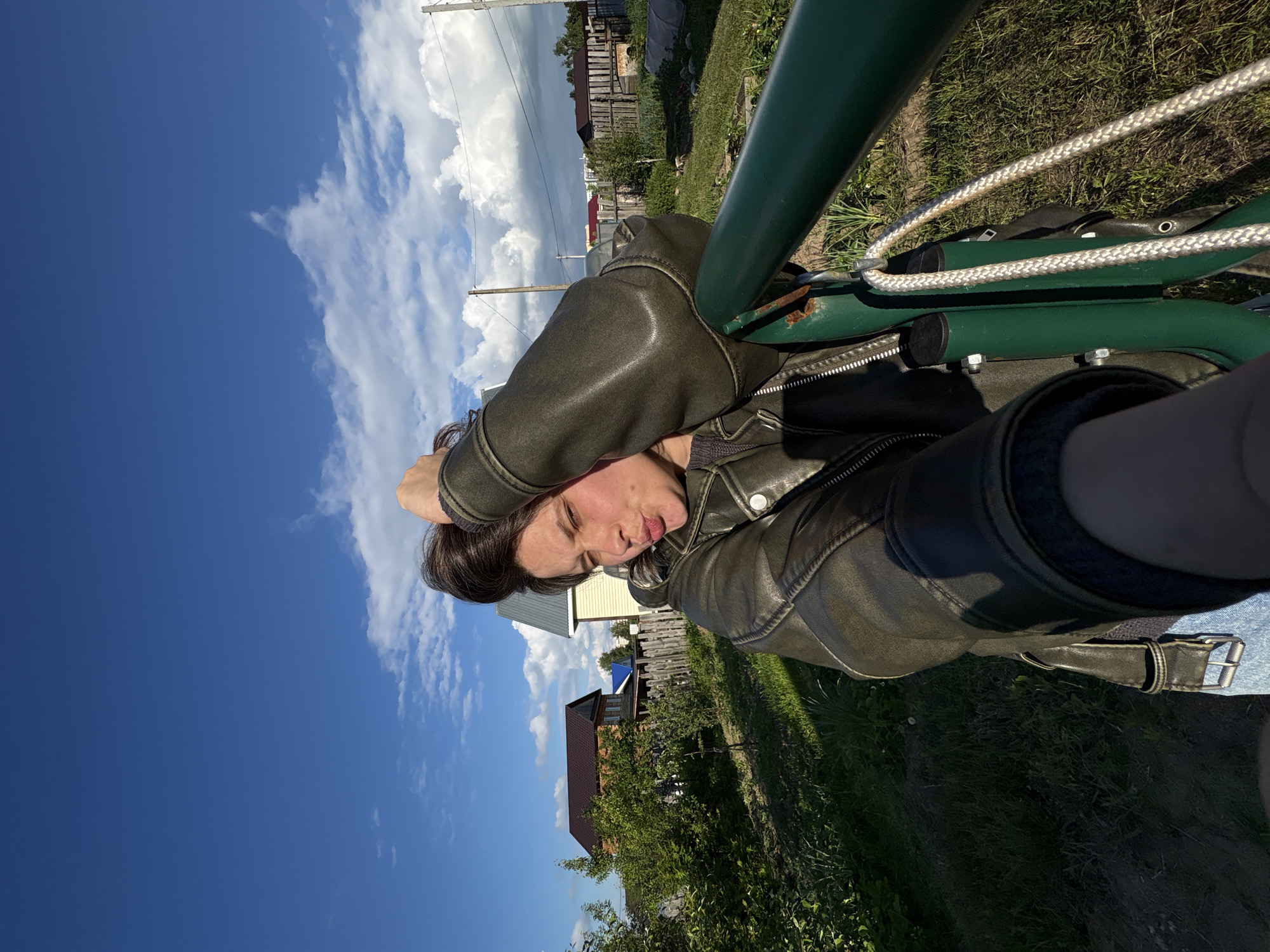 A person is captured in an outdoor, selfie-style shot, taken from a slightly low angle. The subject, a person with dark hair, is positioned in the foreground, wearing a dark olive green leather jacket with visible zippers. Their hands are placed behind or on top of their head, and they are looking towards the camera with a playful, slightly pouting expression.
They appear to be leaning or resting against a bright green metal structure, possibly part of a swing, hammock stand, or outdoor furniture, which occupies a significant portion of the right side of the frame. White braided ropes are looped around this green structure.
The background reveals a bright, sunny day with a deep blue sky adorned with white, fluffy cumulus clouds. In the middle ground, a rural or village setting is visible, featuring various houses, some with wooden fences, surrounded by green grass and trees. Utility poles and power lines are also present, indicating an inhabited area. The strong sunlight casts clear shadows on the ground, suggesting it is daytime, possibly late morning or afternoon.
The overall scene depicts a relaxed moment in an outdoor, village environment, specifically in Chaikovsky, Russia. No visible text is present in the image.
