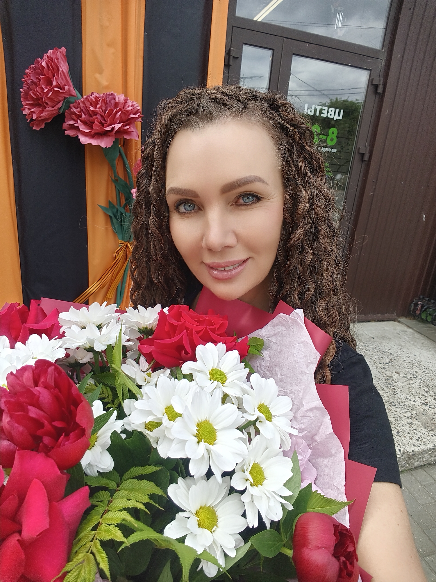 A woman with long, curly, brown hair, styled in braids, is taking a selfie outdoors. She is holding a bouquet of flowers, which includes red roses, red peonies, and white daisies with yellow centers. The woman is wearing a black top. Behind her, a building with brown siding and a dark door with glass panels is visible. A sign with white text, which appears to be in Cyrillic script, is visible on the glass door. The background also features a vertical orange and black striped curtain or banner, and two large red carnation-like artificial flowers. The ground in front of the building is paved with grey concrete. The overall setting suggests an outdoor commercial area.