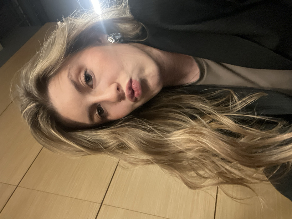 A young woman with long, wavy blonde hair puckers her lips toward the camera. She has fair skin, light eyes, and wears a dark blazer over a tan top. A silver earring is visible in her left ear. The photo is taken at an unusual angle, slightly tilted, with a bright overhead light source. A tiled wall appears to be behind her.