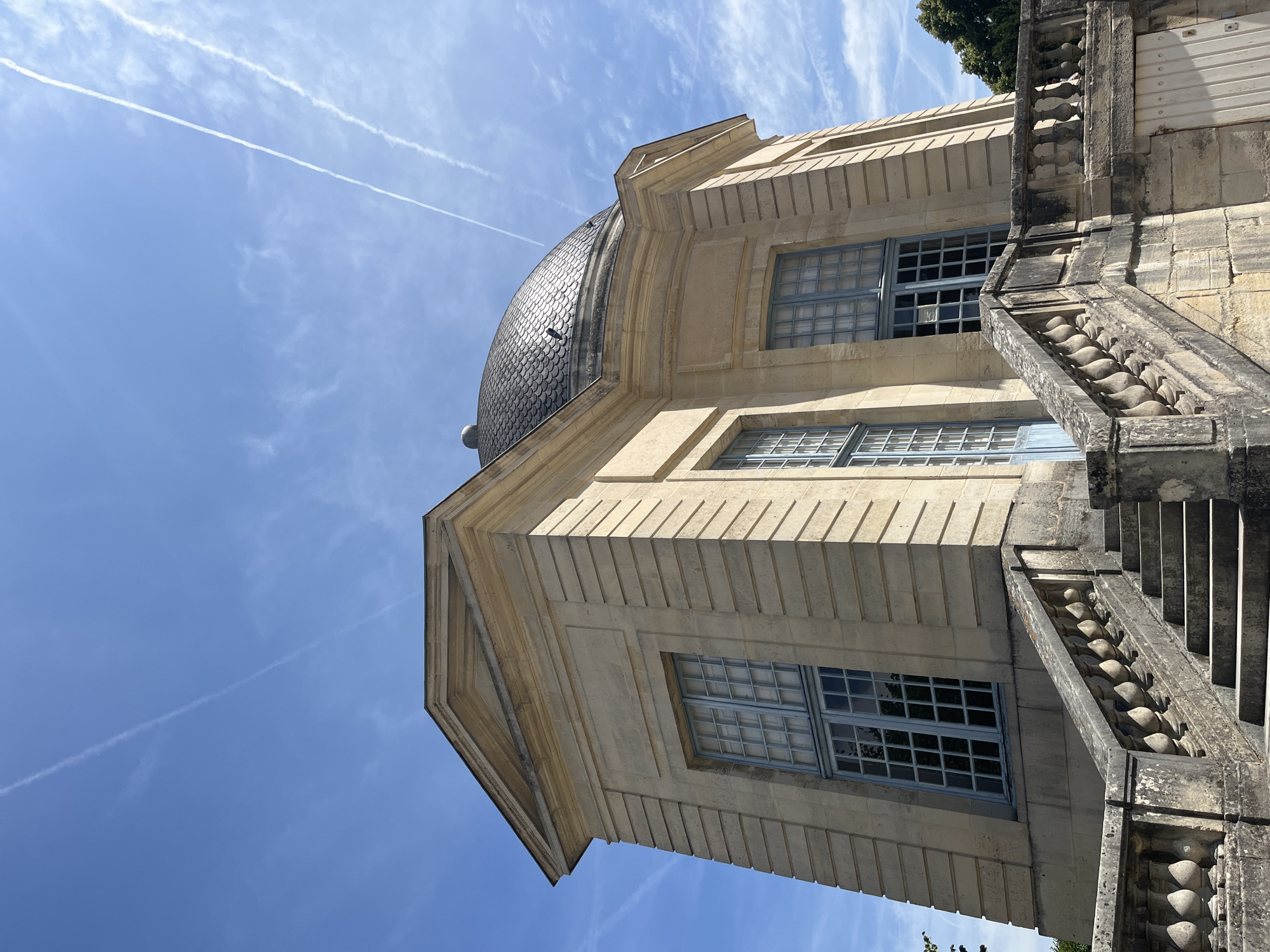 The image shows a low-angle view of a historical stone building with a domed roof, situated in the city of Sceaux, France. The building is made of light-colored stone blocks, with decorative cornices and fluted pilasters. It features large, multi-paned windows with pale blue frames and a matching blue door. A stone balustrade lines a set of stairs leading up to the building. The sky above is clear blue with thin white clouds and contrails. The overall scene conveys a sense of grandeur and historical significance.