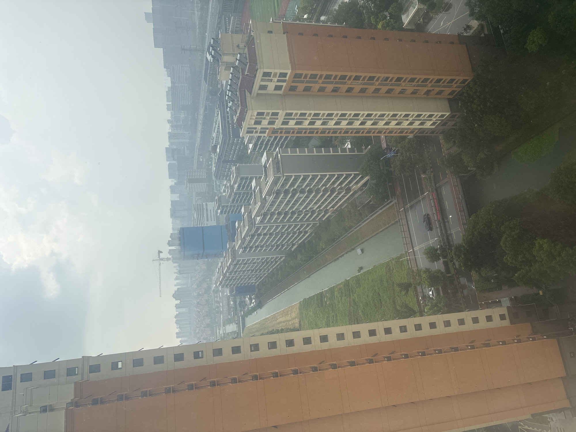 The image shows an aerial view of a residential area in Suzhou Industrial Park, China, characterized by high-rise apartment buildings lining a canal. The foreground is dominated by a tall, orange-hued apartment building on the left, with its facade featuring a grid of dark windows and balconies. To the right of this building, a narrow canal flows, bordered by a strip of green grass and vegetation on one side and a row of taller, grey apartment buildings on the other. A road runs parallel to the canal, crossed by a bridge where two cars are visible. Further into the background, more apartment blocks and urban structures stretch towards a hazy horizon, interspersed with construction cranes and what appears to be a sports field. The sky is overcast, suggesting a muted or hazy daylight.