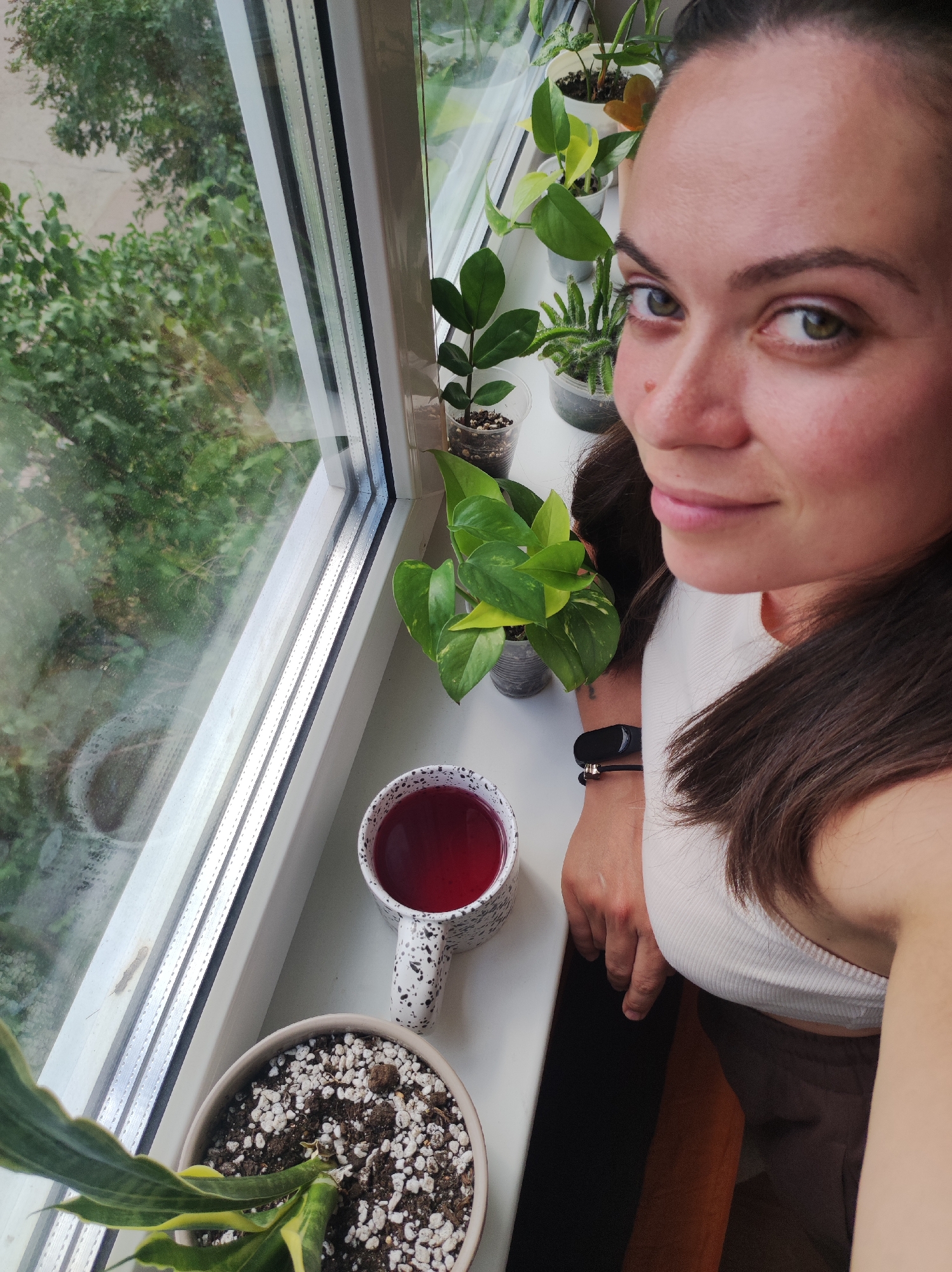 A woman with dark hair and green eyes is taking a selfie indoors, by a window. She is wearing a white tank top and a black fitness tracker on her left wrist. In front of her, on the windowsill, are several potted plants and a mug filled with a red liquid. The window overlooks greenery, suggesting an outdoor environment.