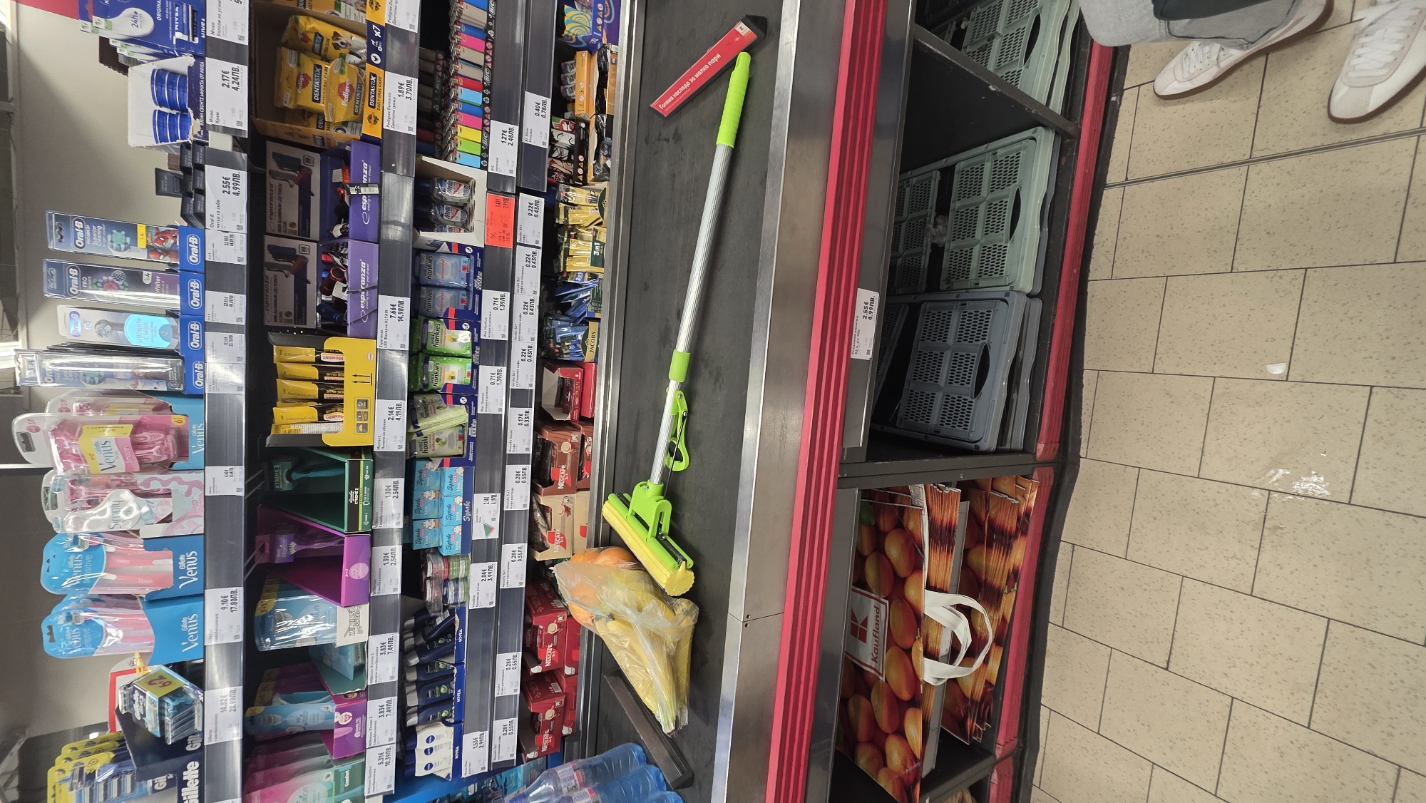 The image shows a scene in a grocery store, likely in Blagoevgrad, Bulgaria. A checkout conveyor belt is visible with various items, including a green and silver sponge mop, a bag of oranges and bananas, and bottles of water. Adjacent to the belt are shelves stocked with products like razors (Venus, Gillette), toothbrushes (Oral-B), cosmetics (Nivea), and candies (Dentastix). Additionally, there are Kaufland shopping bags below the checkout area. A person wearing white sneakers is seen in the upper right corner. Prices in Bulgarian Levs and Euros are displayed on shelf labels.