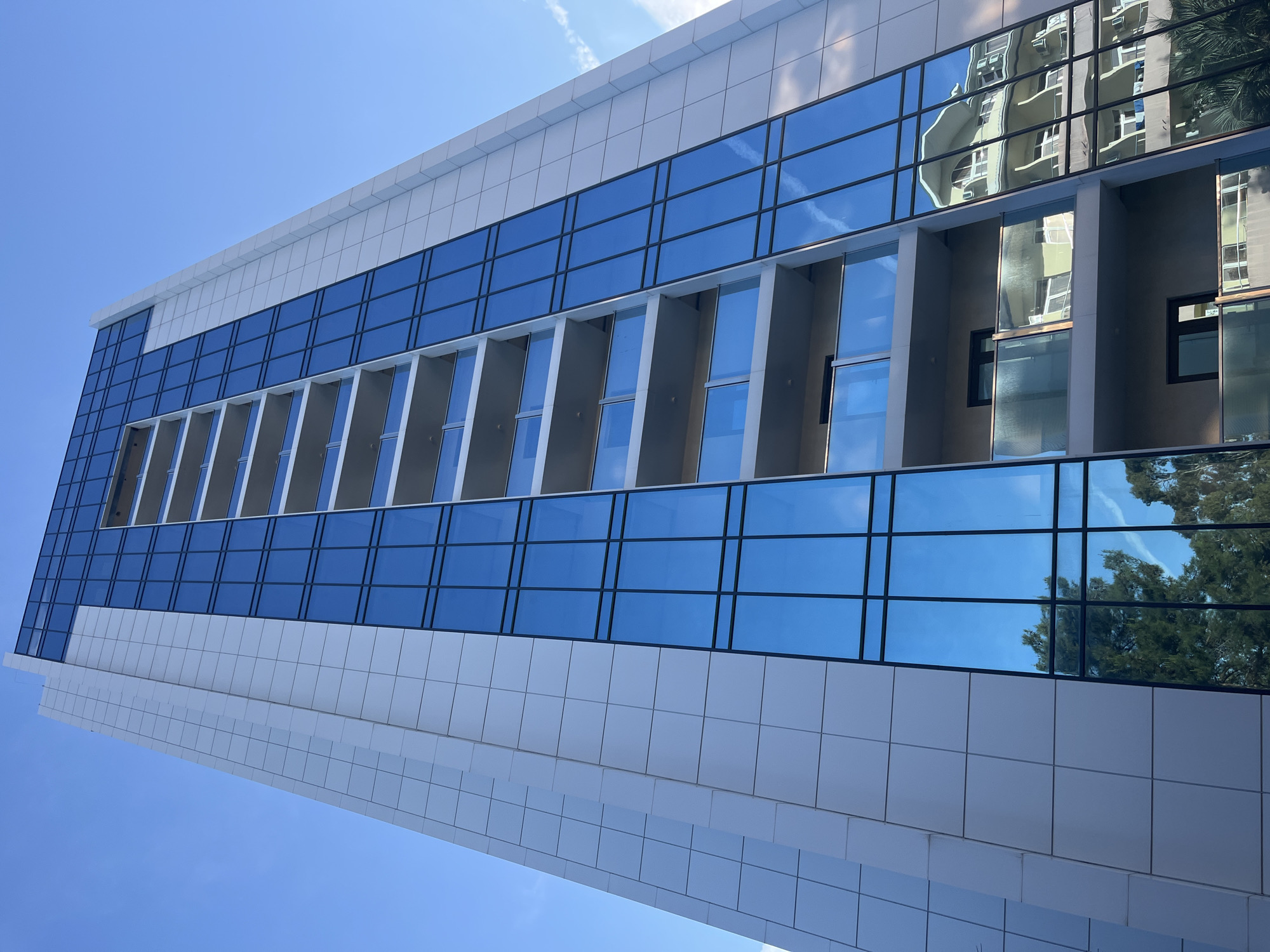 A low-angle, full-frame, vertical, close-up shot captures the facade of a modern, tall, residential building with a clear blue sky overhead, likely in Sochi, Russia. The building's exterior is a striking combination of white paneling and large, reflective blue glass windows. The white panels are arranged in a grid pattern, creating a textured and geometric surface. The blue glass windows are also sectioned into a grid, with thin black frames.
The building features multiple balconies with clear glass railings and white or light-colored overhangs, creating horizontal bands across the facade. These balconies are stacked vertically, emphasizing the building's height. The reflective glass mirrors the sky, clouds, and the surrounding environment, including the lush green foliage of trees and the architecture of a building across the street, which appears to be older and more ornate with curved balconies and arched windows. The reflection is distorted due to the building's angles, adding a dynamic element to the image.
The lighting suggests it is daytime, with the sun casting a bright light that accentuates the building's features and the reflectivity of the windows. No people are visible in the image. The overall impression is one of contemporary urban architecture, with a focus on clean lines and a blend of solid and transparent materials.