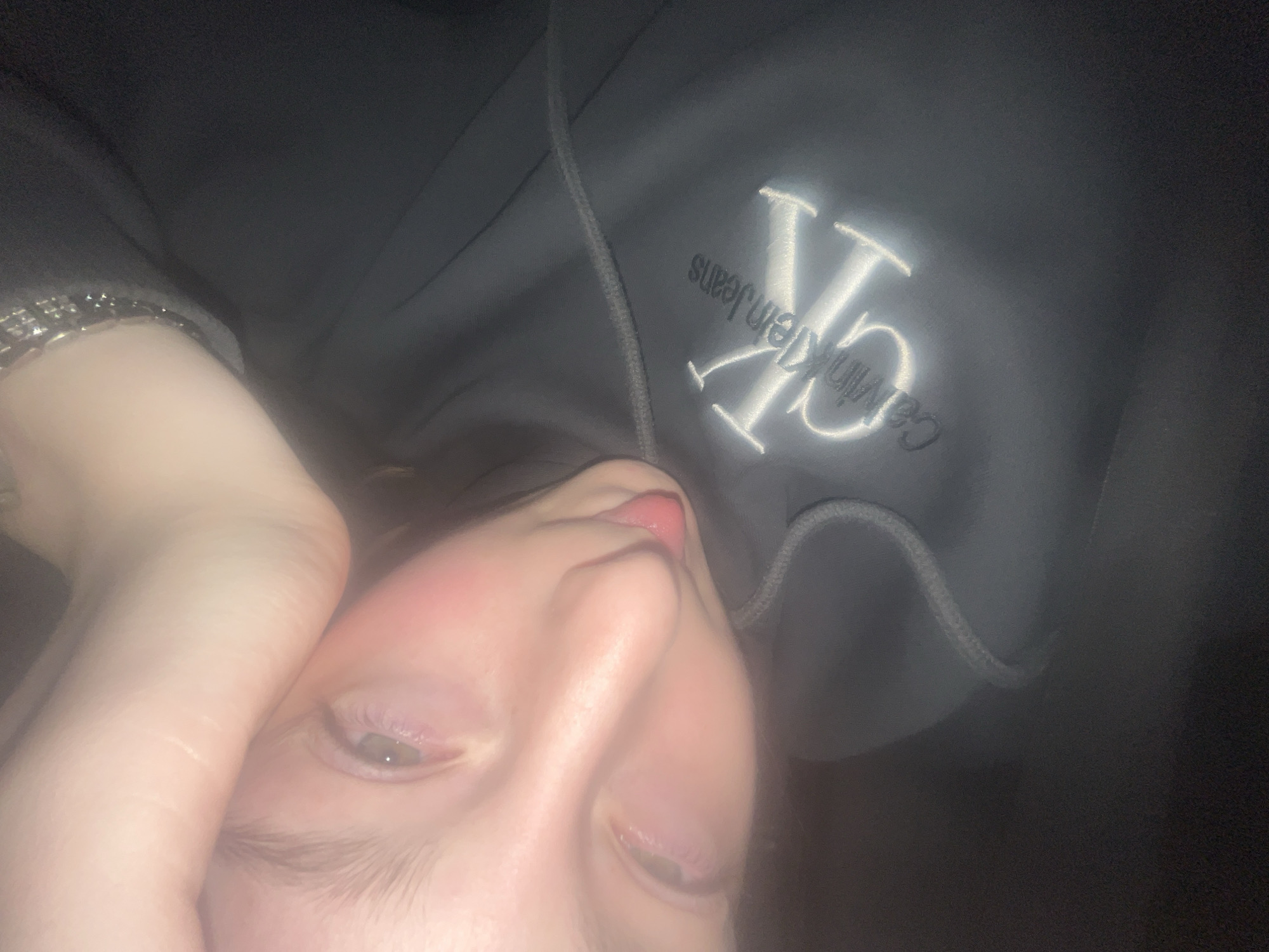 A low-light, rotated close-up portrait shows a person's face and upper body from a worm's-eye view. The person appears to be lying down, looking upwards towards the camera, with their head tilted to the right. Their right arm is visible, partly obscuring the left side of their face, and a metallic bracelet adorns their wrist. The person has fair skin, with a slight flush on the cheeks, and their lips are parted with the tip of their tongue subtly showing. One eye is partially visible, appearing to be light-colored.
The person is wearing a dark gray hoodie, clearly visible in the upper portion of the image. On the front of the hoodie, there is a prominent embroidered logo in white or light-colored thread, featuring the intertwined "CK" symbol. Below and slightly to the left of the "CK" logo, the text "Calvin Klein Jeans" is embroidered horizontally in a smaller font. The hoodie also shows a visible drawstring on the right side.
The background is dark and indistinct, suggesting an indoor setting with low light conditions. The image has a soft focus, giving it a somewhat hazy or ethereal quality. The overall mood is casual and intimate. The location context provided is Varna, Bulgaria.