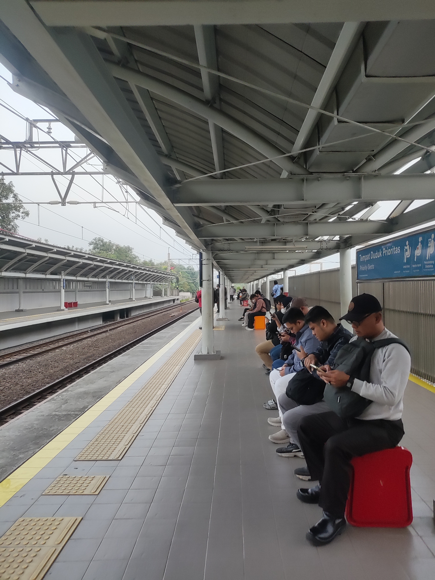 The image depicts a train platform in Telagamurni, Indonesia. Several people are seated on a bench on the right side of the platform, many of them looking at their mobile phones. A red luggage-like object is placed on the ground in front of one of the seated individuals. The platform is covered by a metal roof supported by pillars. Adjacent to the platform are train tracks. The environment appears to be an outdoor train station with a cloudy sky. A sign with the text "Tempat Duduk Prioritas" and "Priority Seats" is visible on a blue panel.