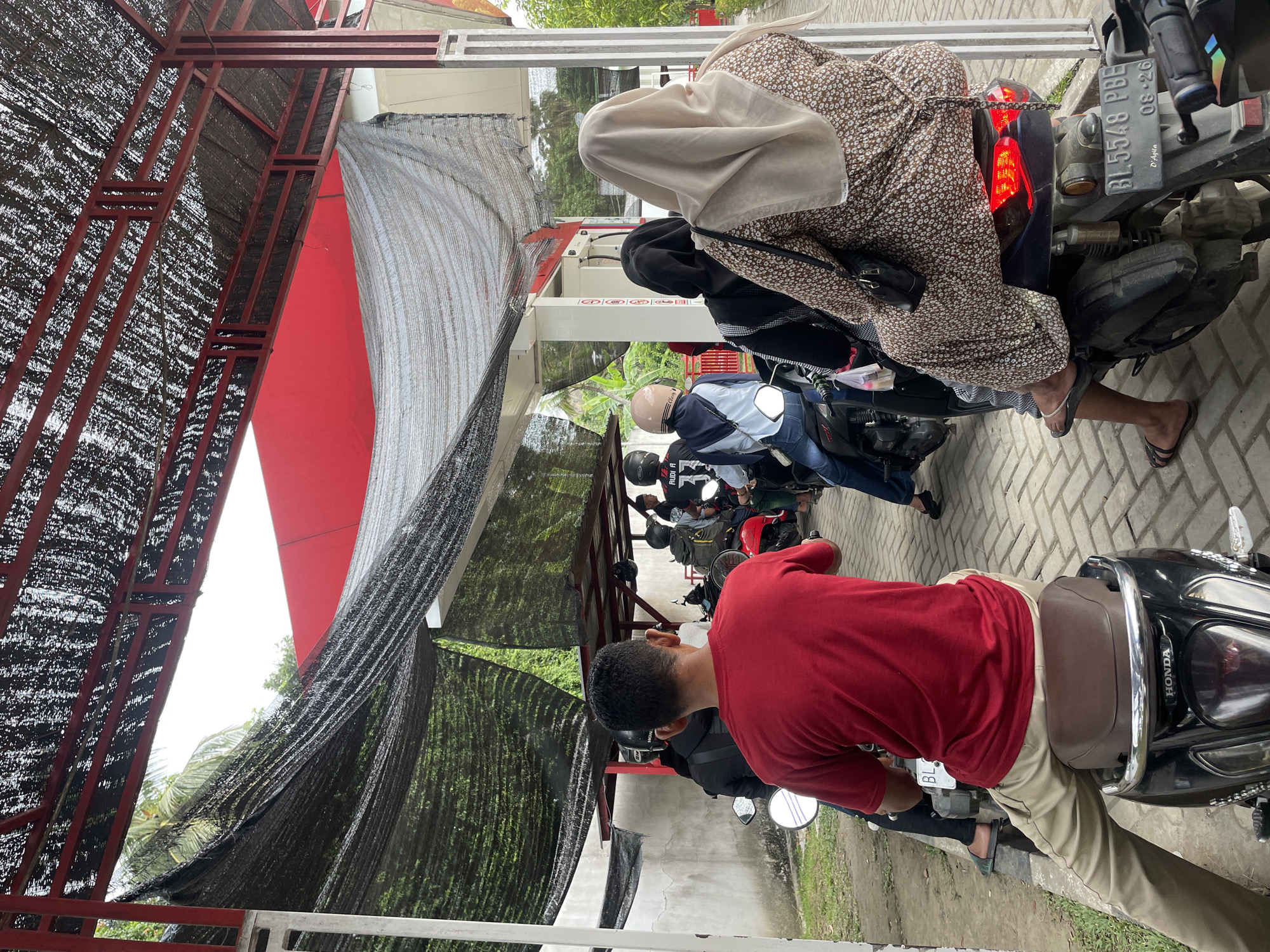 The image depicts a line of motorcycles with riders waiting under a covered structure, likely a gas station canopy, in Glee Gogo, Indonesia. A man in a red shirt is in the foreground, sitting on a black Honda scooter. Behind him, several other individuals are on their motorcycles, some wearing helmets. The structure's roof is made of dark netting stretched over a red metal frame. To the left, a concrete wall is visible, and to the right, part of a building with red accents is present. The ground is paved with interlocking bricks. The overall impression is of a typical scene at a local service station during a potentially busy period.