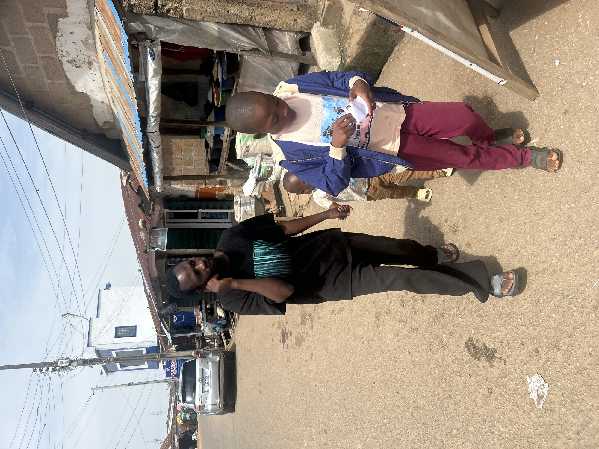 The image depicts a street scene in Jos, Nigeria, with two young boys in the foreground. The boy on the left is wearing a black t-shirt and dark pants, while the boy on the right is wearing a light-colored t-shirt with a graphic print and a blue jacket over it, paired with maroon pants. The boy on the right is holding a small white object, possibly a piece of paper.
Behind the children, there are structures that appear to be makeshift stalls or shops, with corrugated metal roofs and brick walls. Bags of what look like cement or other materials are stacked nearby. In the background, a silver SUV is parked on the asphalt road, and electrical wires are strung overhead. A building with white and blue accents is also visible in the distance.
The ground is a paved road, and shadows suggest it is daytime, likely sunny. The overall scene gives the impression of a busy, outdoor market or street environment. There is no text clearly legible in the image.