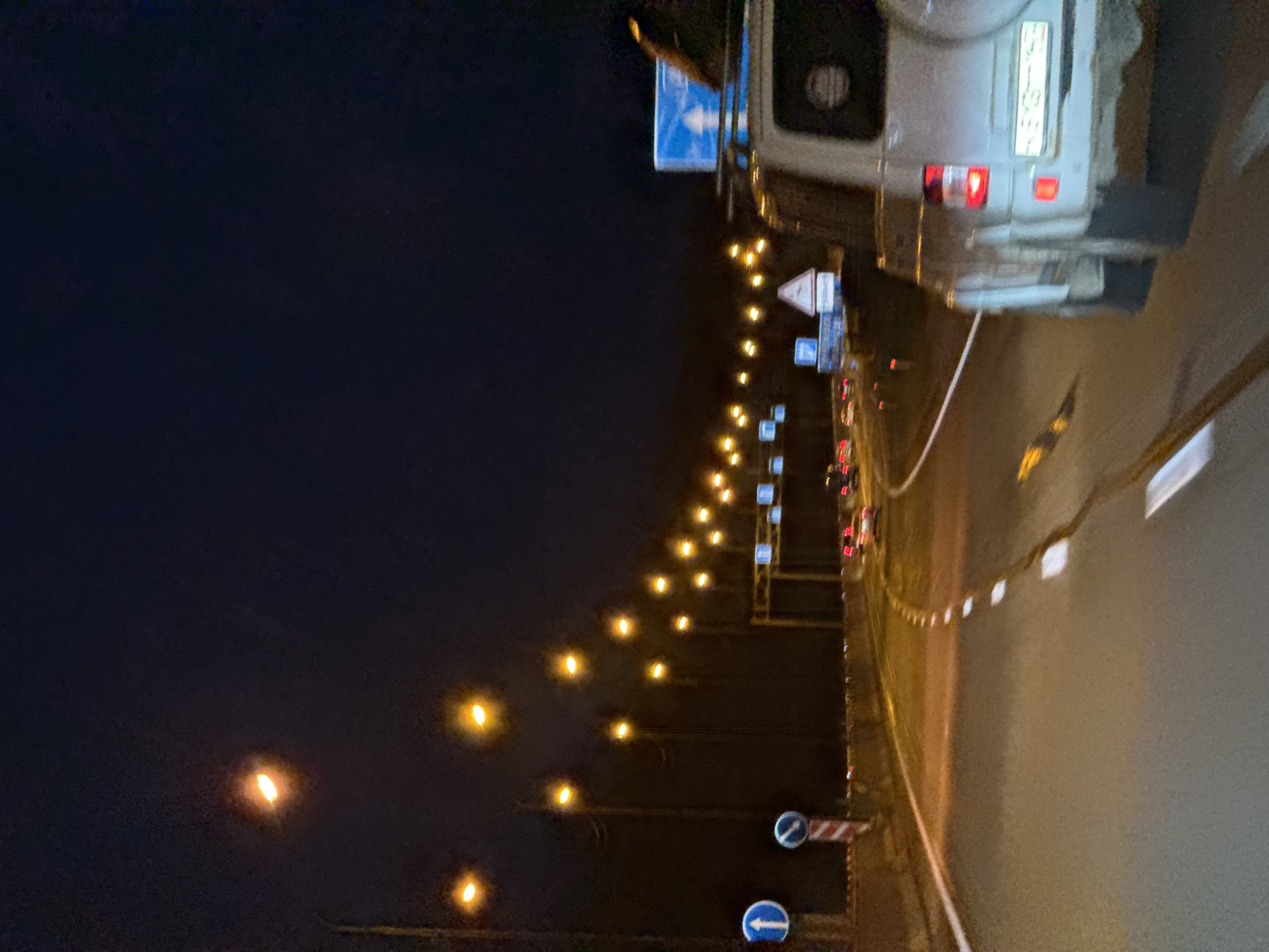 The image depicts a nighttime scene on a highway or main road. Streetlights illuminate the road and the dark, clear sky. Several cars are visible, including a white SUV in the foreground on the right, appearing to be moving forward. Other vehicles are seen further down the road, their taillights glowing red. Traffic signs are present, including directional arrows and a triangular warning sign. The road itself has white lane markings. The environment appears to be outside of a city, possibly rural or suburban, given the presence of trees lining the road in the distance. The location is identified as Vaskelovo, Russia.