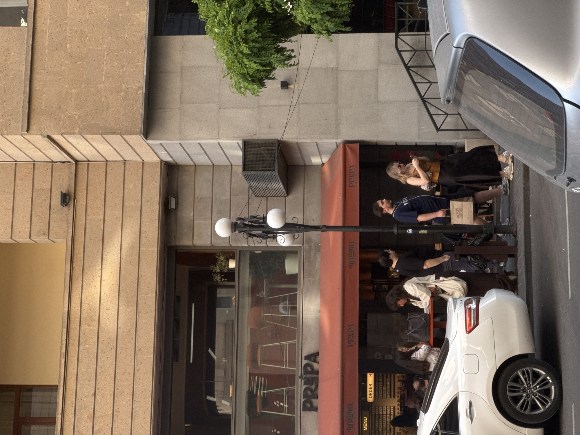 This image shows the exterior of "PREPA," a cafe in Yerevan, Armenia. Several people are sitting at tables outside the cafe, some are eating and drinking. A street lamp stands in front of the cafe. Parked cars are visible on the street. The building is made of stone and has a large awning above the outdoor seating area.