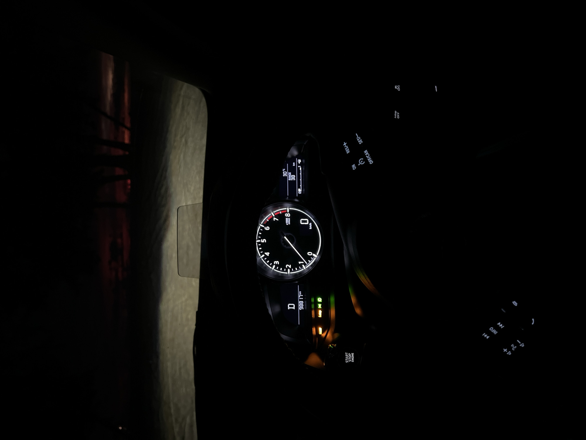The image depicts the interior of a car at night, looking forward through the windshield. The dashboard displays are illuminated, showing a digital readout of "D" for drive and "98817 km" for mileage. The tachometer is also visible, indicating 0 RPM. To the right, a temperature display shows "30°C" and a range of "38 km." A head-up display projects information onto the windshield. Outside, the road ahead is illuminated by headlights, and a dark, silhouetted treeline is visible against a dimly lit, reddish sky, suggesting dusk or dawn. The environment appears to be an outdoor, possibly rural setting due to the treeline and lack of visible city structures. The image is dark overall, with the primary light sources being the car's displays and headlights.