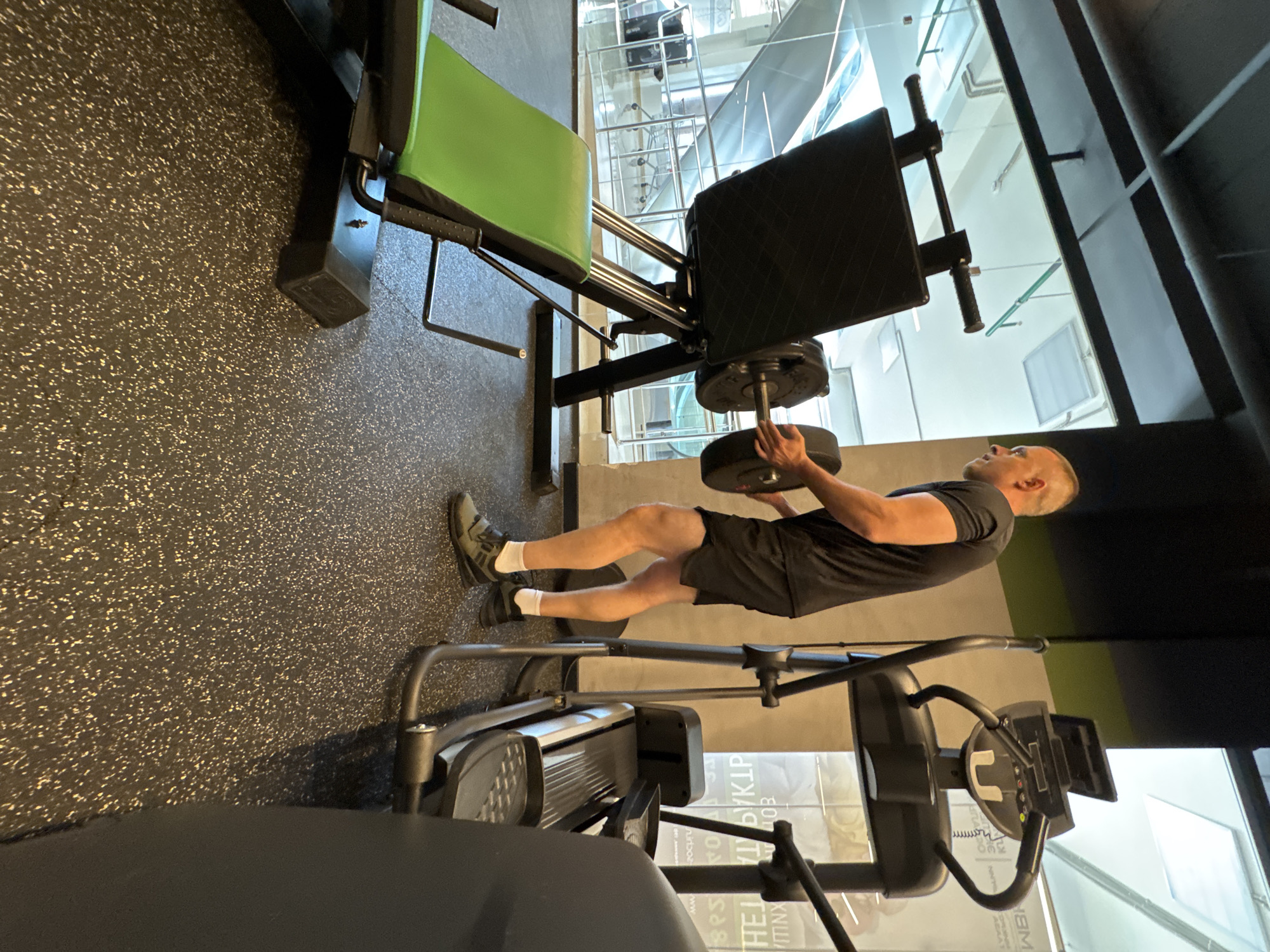 A man with a shaved head, wearing a black t-shirt and black shorts, is performing a leg press exercise at a gym. He is positioned on a leg press machine with a green seat and a black padded backrest. His feet are flat against the weight plate, and his knees are bent. He is holding onto the handles of the machine. In the background, a large window reveals a bright, possibly daytime view of an urban or industrial setting with multiple levels and metal structures. The gym floor is a speckled black rubber mat. Visible text on a poster in the background is partially obscured but appears to include Cyrillic script. The overall scene suggests an indoor workout session in a well-equipped fitness facility.