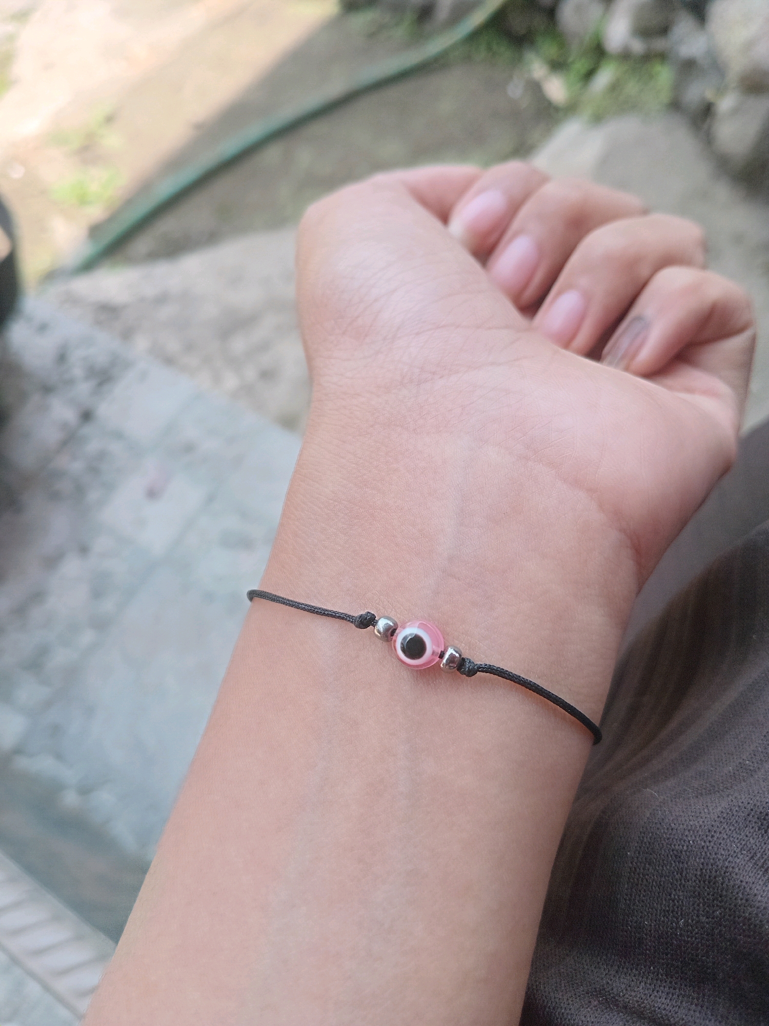 A close-up shot shows a person's wrist wearing a thin black cord bracelet with a pink evil eye charm. The charm has a black pupil and a white iris, outlined in pink. Two small silver beads flank the charm. The background is out of focus, showing an outdoor scene with concrete and greenery, suggesting a casual outdoor setting potentially in Claket, Indonesia, given the context.