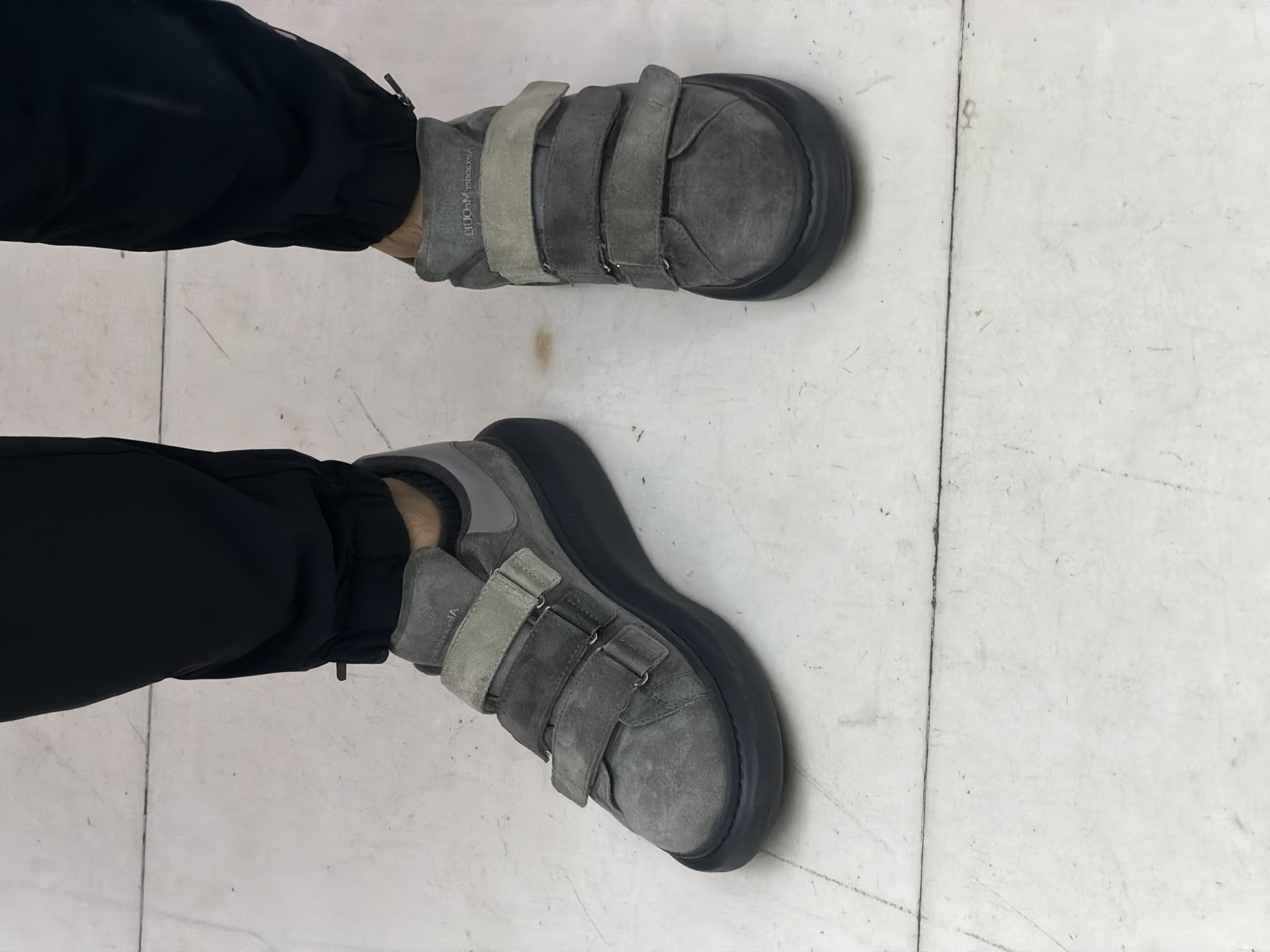A top-down view depicts a person wearing gray suede Alexander McQueen sneakers with three velcro straps on a light-colored, possibly white, tiled floor. The person is also wearing black pants that cinch at the ankles. The environment appears to be indoors, indicated by the tiled flooring. The city of Santa Clara Ocoyucan, Mexico, cannot be verified from this image.