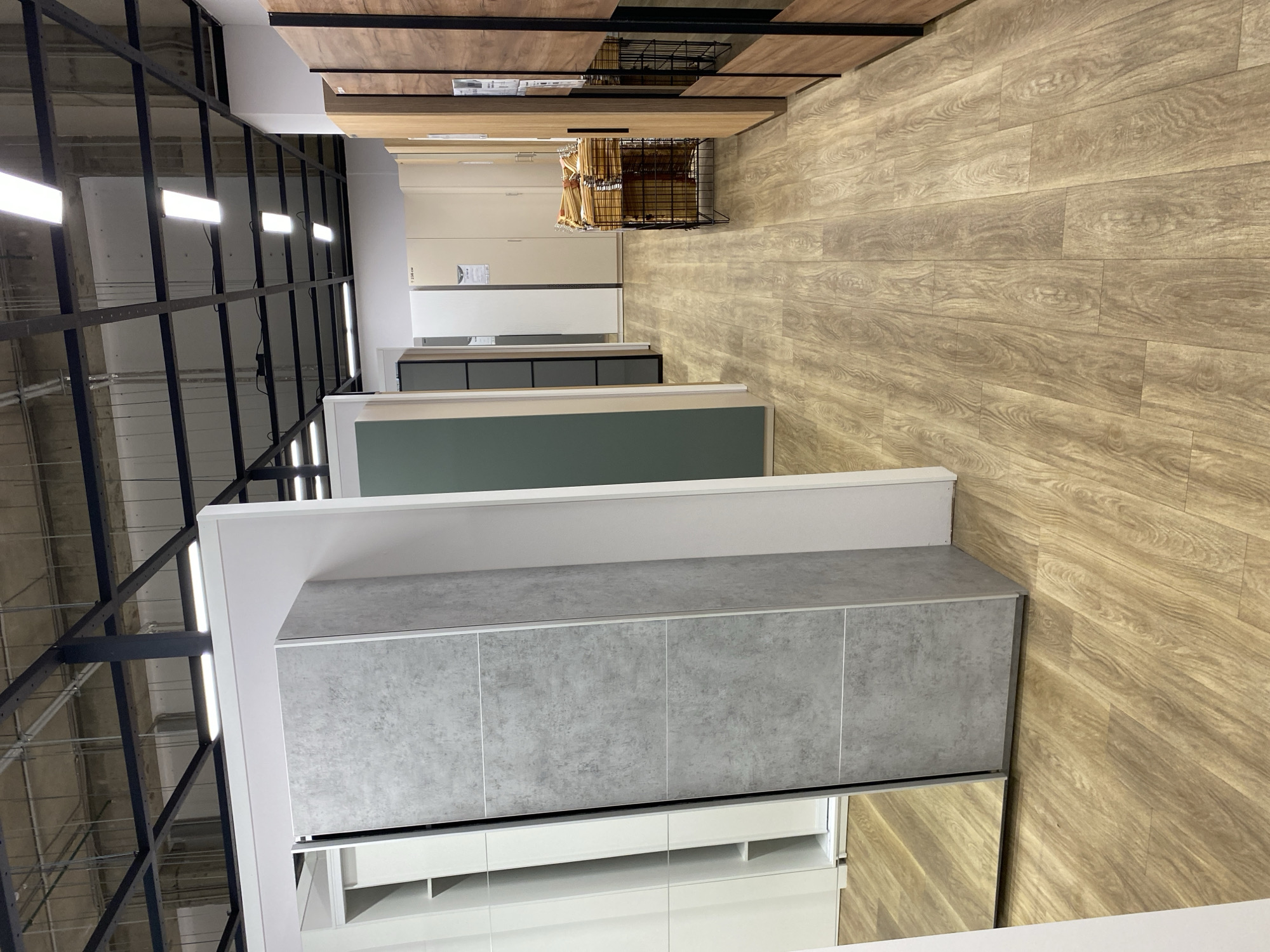 The image shows an indoor setting, likely a showroom or retail space, possibly in the city of Murino, Russia, though this cannot be definitively confirmed from the image alone. The focus is on a display of kitchen or storage units. The units are presented in a receding line of different colors and materials, including grey, green, and white.
The floor is covered with a wood-grain pattern. Above the units, there is a black metal framework or support structure, possibly for lighting or additional displays. Fluorescent lights are attached to the framework, providing illumination.
At the far end of the display, there is a rack with items, possibly kitchen utensils or accessories. Additional shelves or cabinets are visible above.
The scene is devoid of people and actions. It appears to be a static display intended to showcase the available options for kitchen or storage solutions.