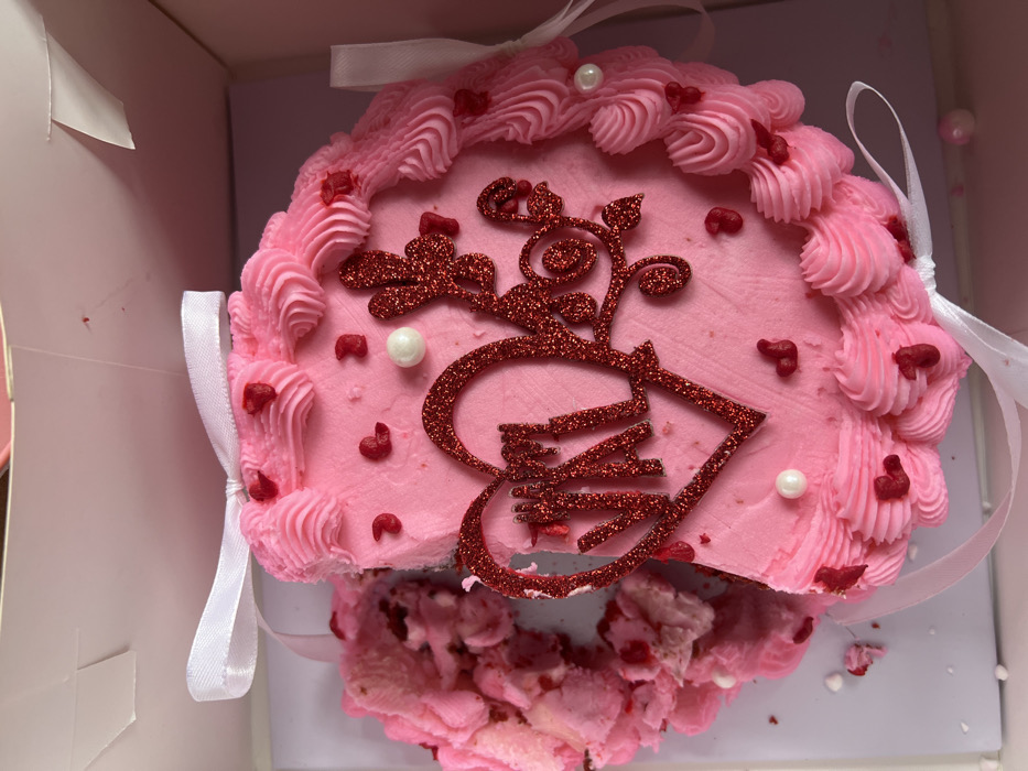 An overhead shot captures a partially eaten or damaged pink cake nestled within a white cardboard box. The cake is predominantly pink, covered in smooth pink frosting, with a ruffled or piped border of lighter pink frosting around its perimeter. Several small, red, heart-shaped sprinkles are scattered across the top and embedded in the piped frosting. White, pearl-like spherical sprinkles are also sparsely placed on the cake's surface.
A large, intricate decoration made of red glitter is centrally placed on the cake. This decoration appears to combine a stylized letter or symbol with flourishes. The lower, wider part of the glitter design seems to incorporate a heart shape with elements resembling piano keys or a keyboard pattern within it. From this base, vine-like or floral flourishes extend upwards and outwards, creating an elegant, ornate design.
The cake shows significant signs of consumption or damage, particularly at the bottom-front edge. A large portion of the cake is missing, and the remaining cake and frosting in that area are heavily disturbed, with crumbs and smeared frosting visible at the bottom of the cake and scattered on the white box surface.
White satin ribbons are tied around the cake, presumably to hold it in place or for decorative purposes, with one bow visible towards the top-left and another ribbon extending from the right side. The interior of the cake box is white cardboard, framing the cake. The overall aesthetic suggests a festive or celebratory occasion, possibly romantic or musical-themed, given the hearts, pink color, and the unique glitter decoration, but the current state of the cake hints at the end of such an event or an unfortunate accident.