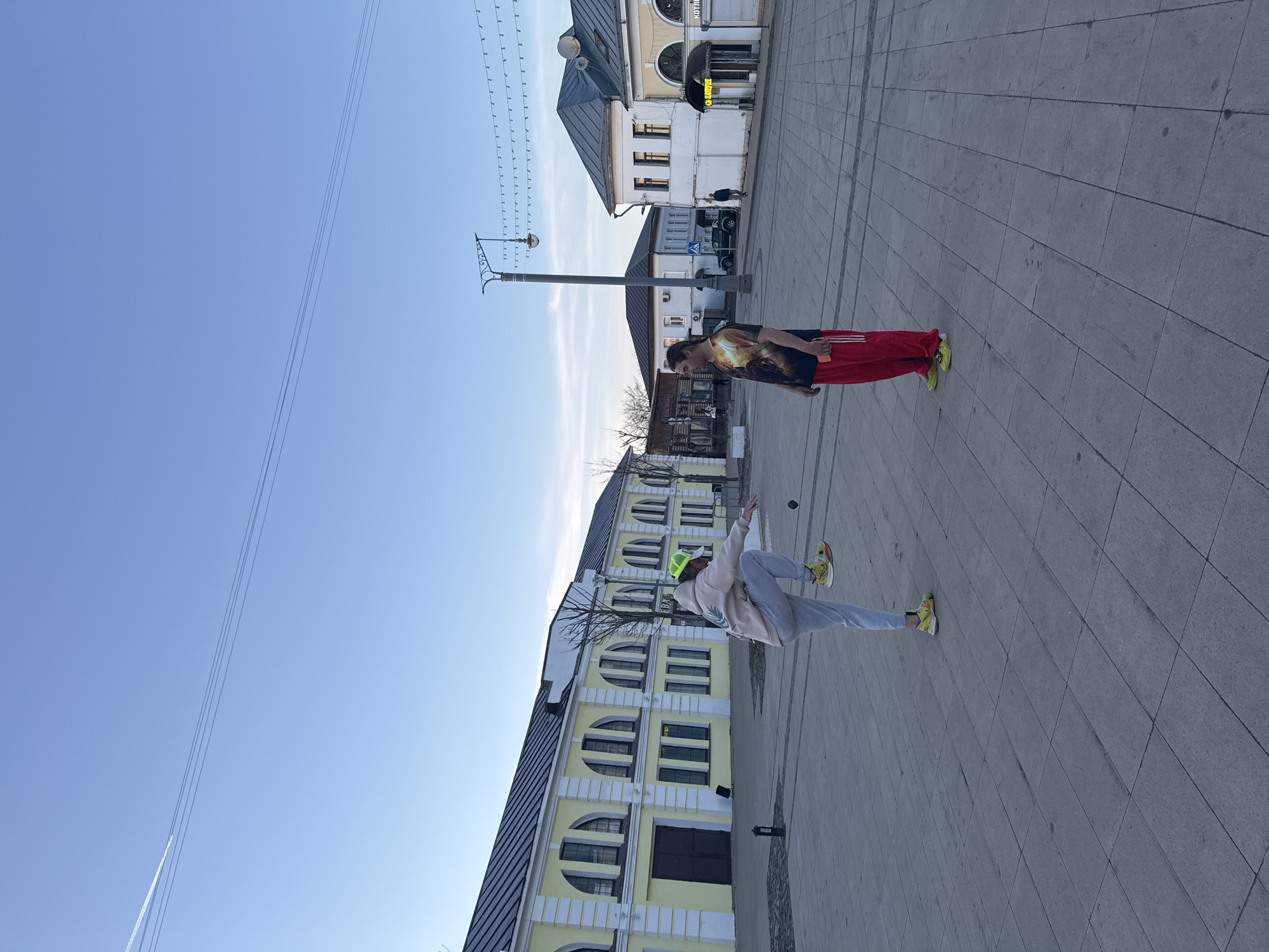 The image captures a street scene in Ново-Никольское, Russia. Two people are visible on a wide, paved pedestrian area. One person, wearing a light-colored hoodie, gray pants, and bright yellow shoes and cap, is mid-action, possibly throwing an object. The other person, wearing a t-shirt with a graphic print and red sweatpants with white stripes, stands still. Background elements include buildings with distinct architectural features, such as arched windows, and overhead power lines.