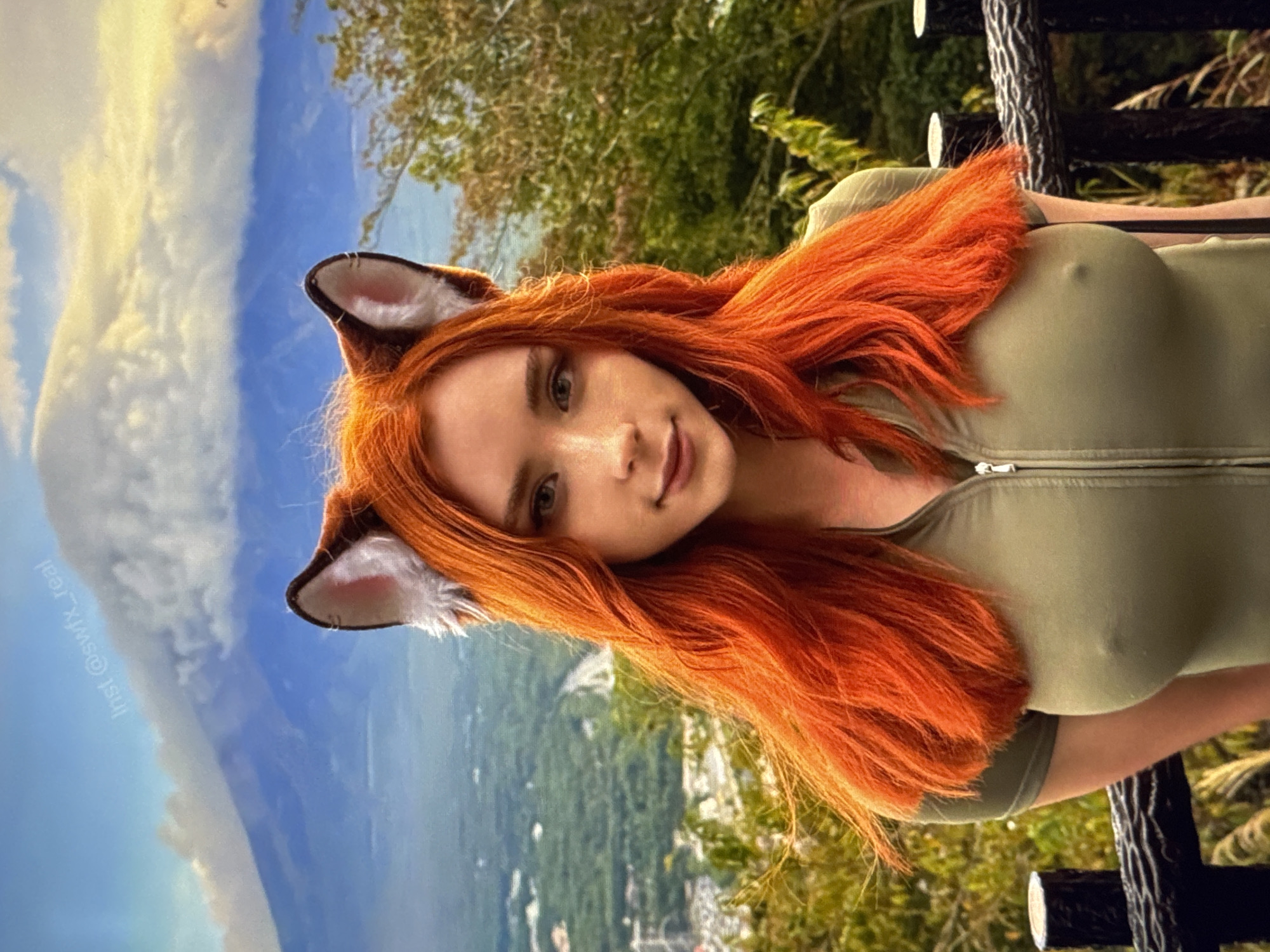 A young woman with bright orange hair and fox ears stands in front of a mountain landscape. She is wearing a green zipped top. The background shows a hazy mountain range under a cloudy sky, with green trees and a village nestled in the valley below. A fence is visible in the foreground.