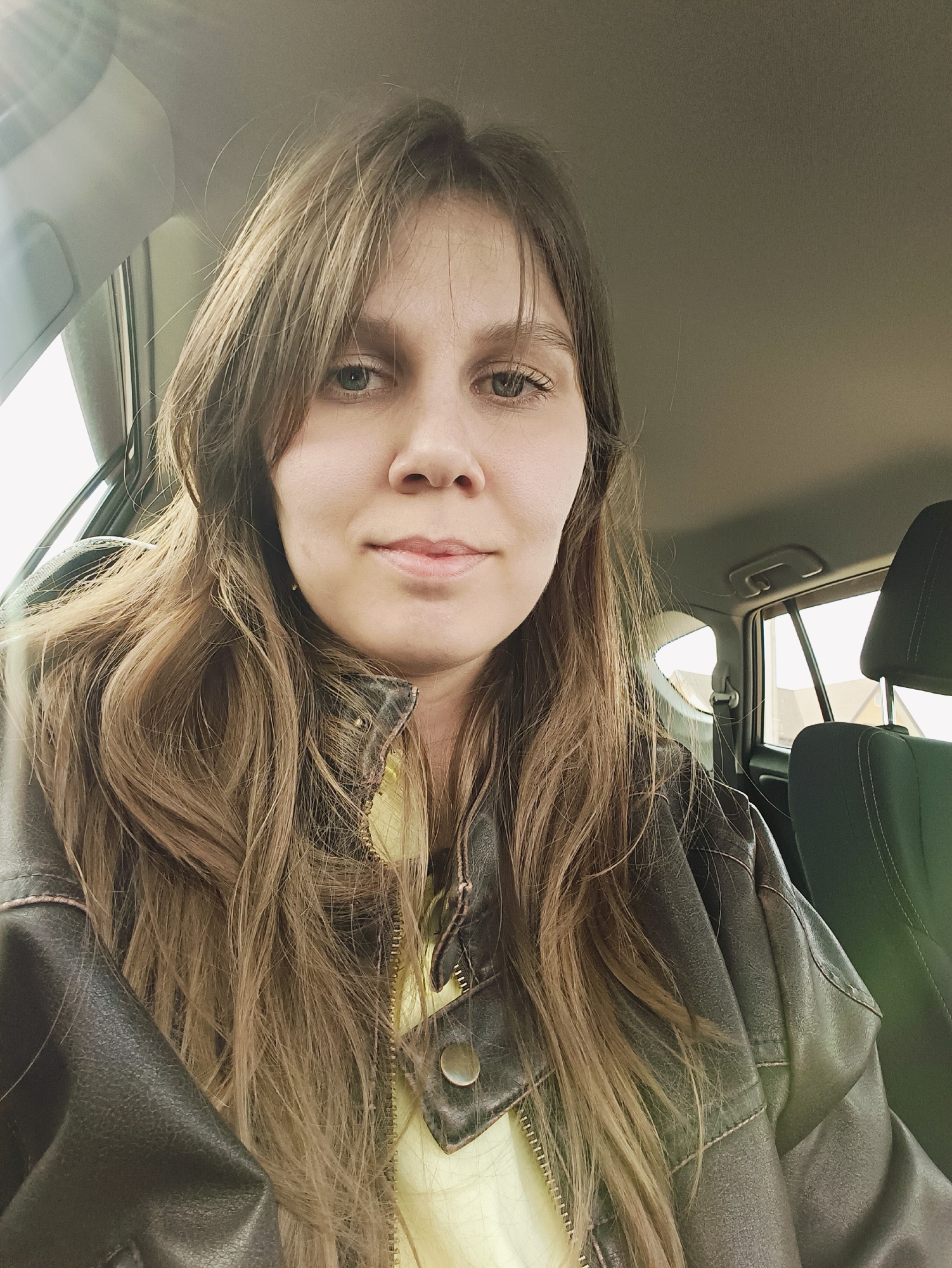 A close-up portrait depicts a female individual seated in the front of a car during daytime. She has long, wavy, light brown hair that cascades over her shoulders and frames her face. Her eyes are light-colored, appearing to be a shade of green or grey-green, and she looks directly at the camera with a neutral to slightly pleasant expression, showing a faint hint of a smile.
She is wearing a dark brown, possibly faux leather, jacket with visible stitching and a zippered front. A light yellow garment is visible underneath her open jacket. The jacket exhibits a textured or worn appearance.
The setting is the interior of a car. Visible car elements include the light-colored headliner/ceiling, parts of the window frames and door pillar, and a dark car seat headrest on the right side of the frame, which appears to be the front passenger headrest. Bright light streams into the vehicle through the windows, creating a noticeable sun glare in the upper left portion of the image, indicating it is daytime. Outside the rear window, a glimpse of an outdoor scene with a light-colored building can be seen. There is no visible text in the media file.
