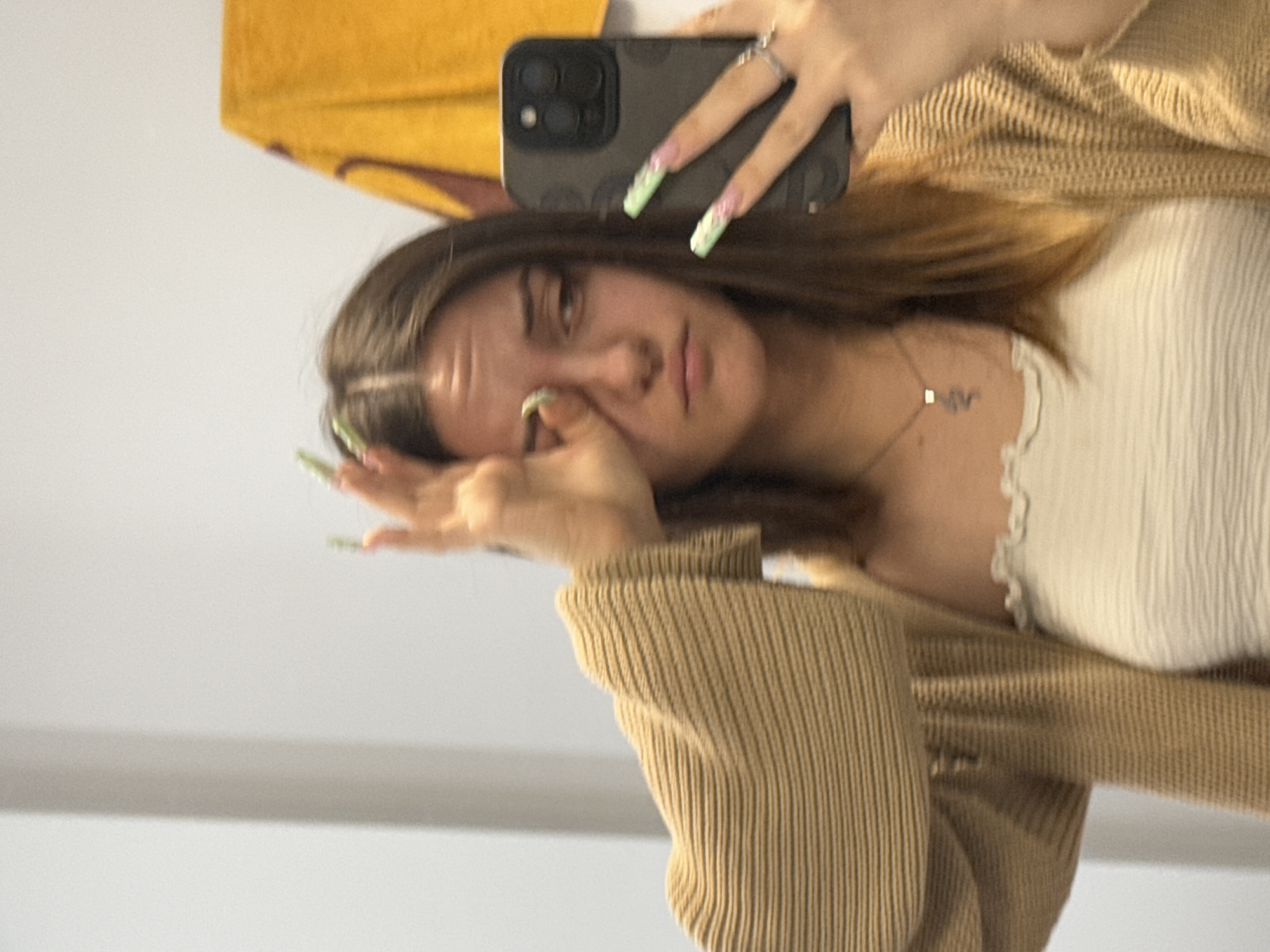 A young woman is taking a selfie in a mirror. She is holding a dark-colored smartphone in her right hand, which is positioned near the top of the frame. She is partially obscuring her face with her left hand, which has long, green-tipped nails. The woman has brown hair with lighter brown highlights and is wearing a white, ribbed top with a frilly edge and a light brown, ribbed cardigan. She also has a thin necklace. In the background, there is a yellow item partially visible in the top left corner. The environment appears to be indoors with white or light-colored walls. There is insufficient information to determine the specific location in Sofia, Bulgaria.