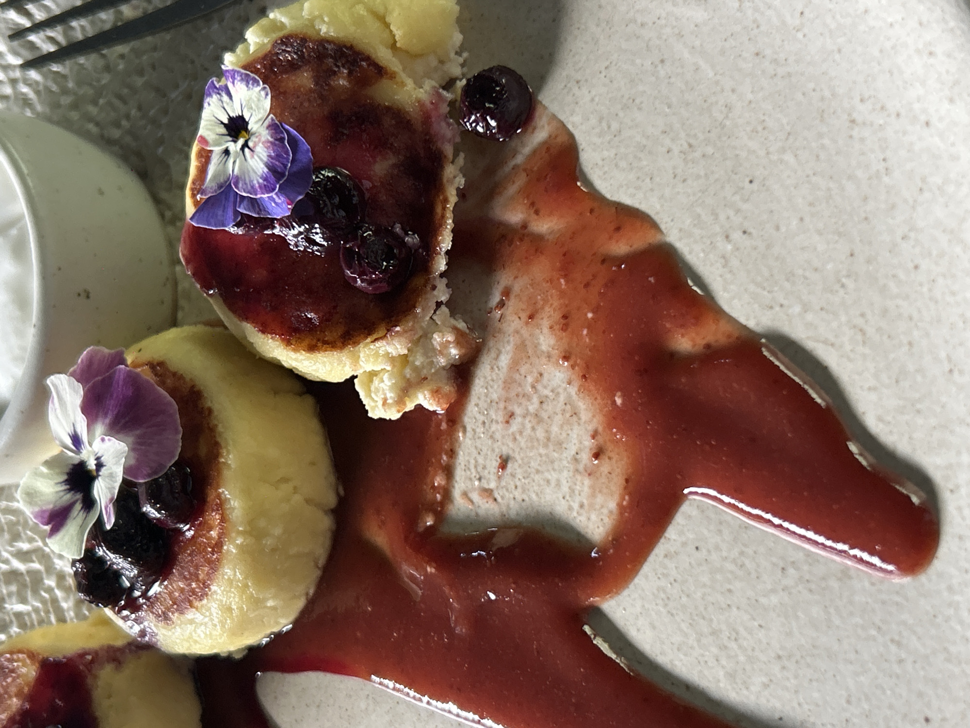 A high-angle, close-up shot shows a plate of what appears to be cottage cheese pancakes, known as Syrniki, served with berry sauce, blueberries, and edible flowers. The pancakes are round and golden-brown, topped with a dark berry sauce and a few whole blueberries. Each is garnished with a small, multi-colored edible flower, possibly a pansy. A generous amount of berry sauce is spread across the light grey, speckled surface of the plate, creating a glossy contrast. Part of a white sauce container and a black fork are visible in the upper left corner. The texture of the pancake suggests a soft, slightly crumbly consistency. The scene suggests a well-presented dish at a restaurant in North Kuta, Indonesia, where this image was reportedly taken.