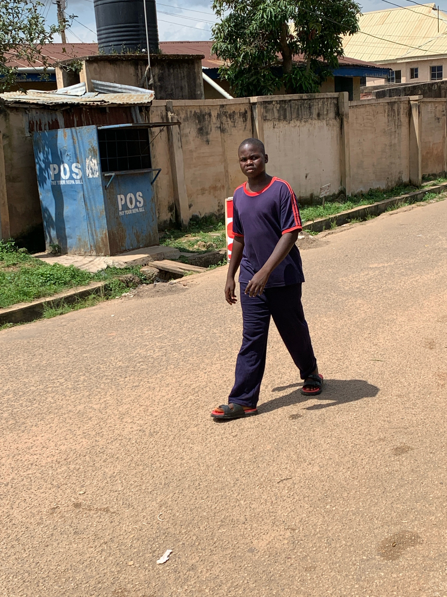 A young boy walks on a dusty road in Jos, Nigeria, casting a long shadow. He wears a dark blue t-shirt with red trim and dark blue track pants, along with black and red sandals. To his left, a blue structure with "POS" and "PAY YOUR NEPA BILL" written on it is visible. This structure appears to be a payment collection point. In the background, a concrete wall and buildings are seen. The scene suggests an outdoor, urban environment.