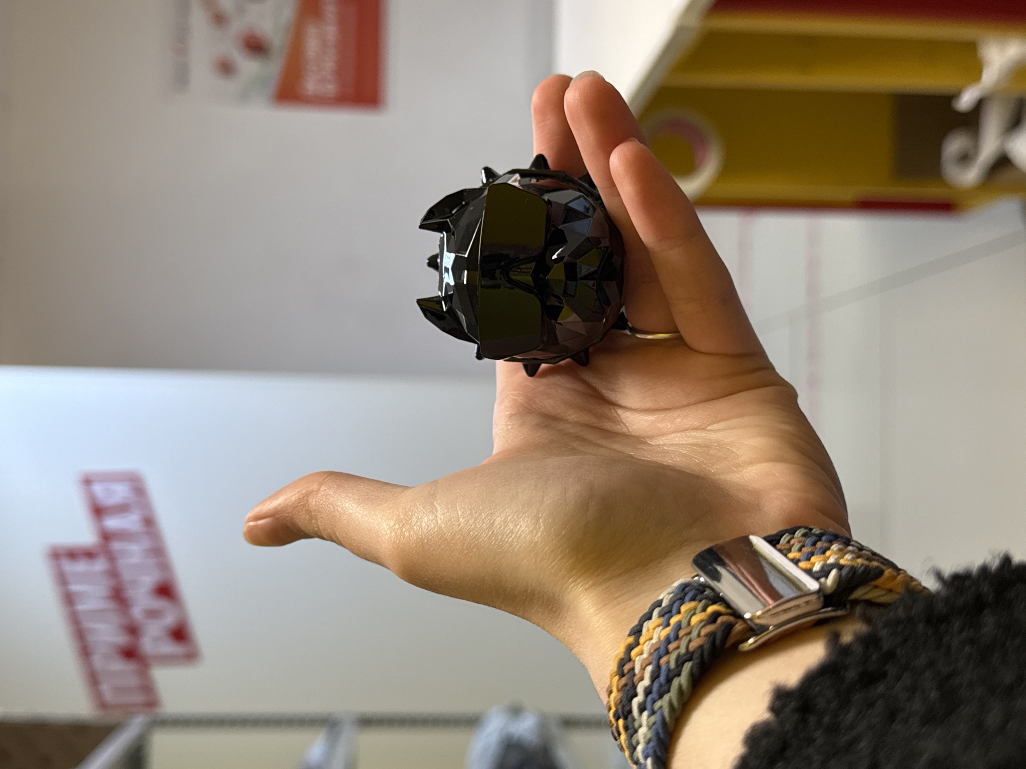 A person's hand holds a small, black, spiked sphere in the foreground. The hand has a gold ring on one finger and a woven bracelet with a metal clasp on the wrist. In the blurred background, there is a white wall with red signage in Cyrillic script, and a glimpse of yellow shelving. The image was taken indoors. Location is not determinable.