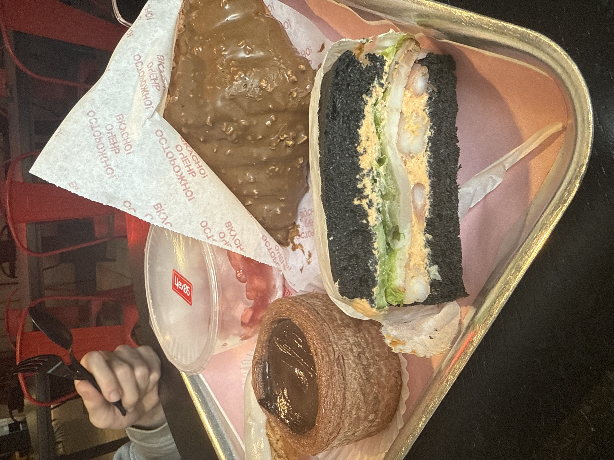 A tray of food sits on a dark surface, likely a table, in Saint Petersburg, Russia. The tray contains a variety of pastries and a sandwich. There is a large chocolate-covered croissant, a smaller chocolate-filled pastry, and a black sandwich with shrimp filling. A clear plastic container labeled "Цех85" (Tsekh85) is also visible. A hand holding a black plastic fork and spoon is positioned near the tray. The packaging includes the text "ВКАСНО ОЛЕНР ОСЛОБОЖНОЇ".