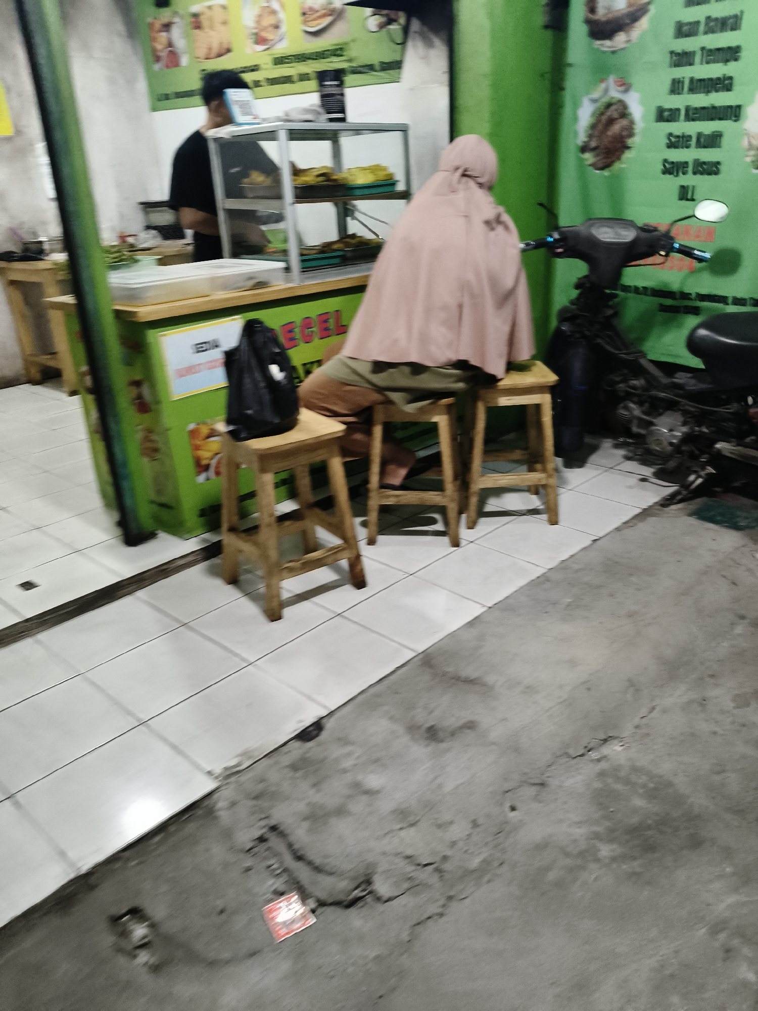 In South Tangerang, Indonesia, a food stall displays a menu featuring items like "Ikan Bawal," "Tahu Tempe," "Ati Ampela," "Ikan Kembung," "Sate Kulit," and "Saye Usus." A person, possibly an employee, wearing a black t-shirt is visible behind a glass display case, while another individual, wearing a light brown hijab and clothing, is seated on a wooden stool. Next to the seated person, a black bag rests on another wooden stool. A motorcycle is parked nearby, and the stall is decorated with green signage. The ground is a combination of white tiles and concrete.