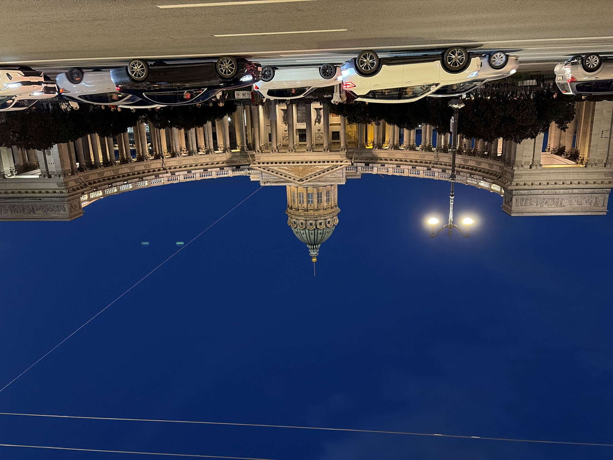 The image shows Kazan Cathedral in Saint Petersburg, Russia, at dusk. The grand neoclassical cathedral, with its iconic colonnade and dome, is illuminated by streetlights. A busy street with several cars driving in different lanes is in the foreground. Some pedestrians can be seen walking on the sidewalk in front of the cathedral. The sky is a deep blue, indicating that it is either early evening or late night. The scene captures the architectural grandeur of the cathedral and the vibrant urban life of Saint Petersburg.