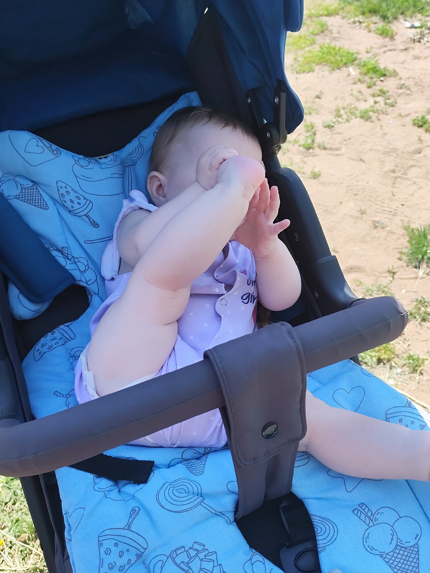 A baby is sitting in a blue stroller with ice cream and lollipop patterns on the fabric. The baby, wearing a purple outfit, has their foot up to their face, obscuring their vision. The stroller has a grey safety bar and straps. The background shows a sandy or dirt area with some sparse grass. The location is the city of Фоки, Russia.